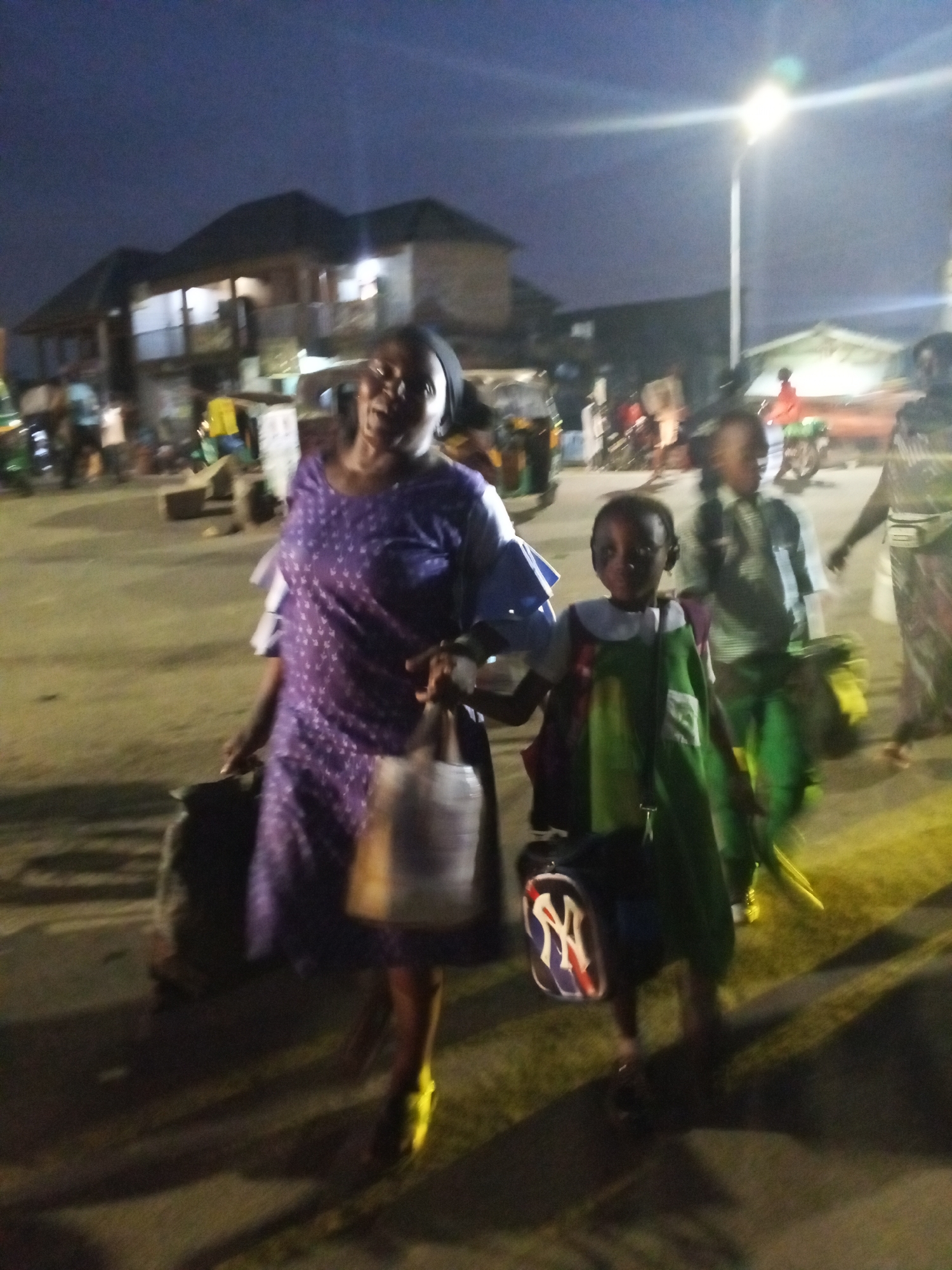 The image captures an outdoor scene in Gwong, Nigeria, at dusk or night, indicated by the low lighting. A woman in a purple dress with white polka dots and ruffled sleeves is walking alongside a child wearing a green school uniform. Each is carrying a bag. The woman's bag is a light color, possibly white or beige, while the child's is a blue bag with a stylized "NY" logo in white with red and blue accents. The woman also appears to be wearing yellow shoes. A street light is visible in the background. Behind them, other people are walking, including another child in a similar school uniform, and motorized vehicles are parked along the side of the road. Buildings are visible in the background, suggesting a residential or commercial area.