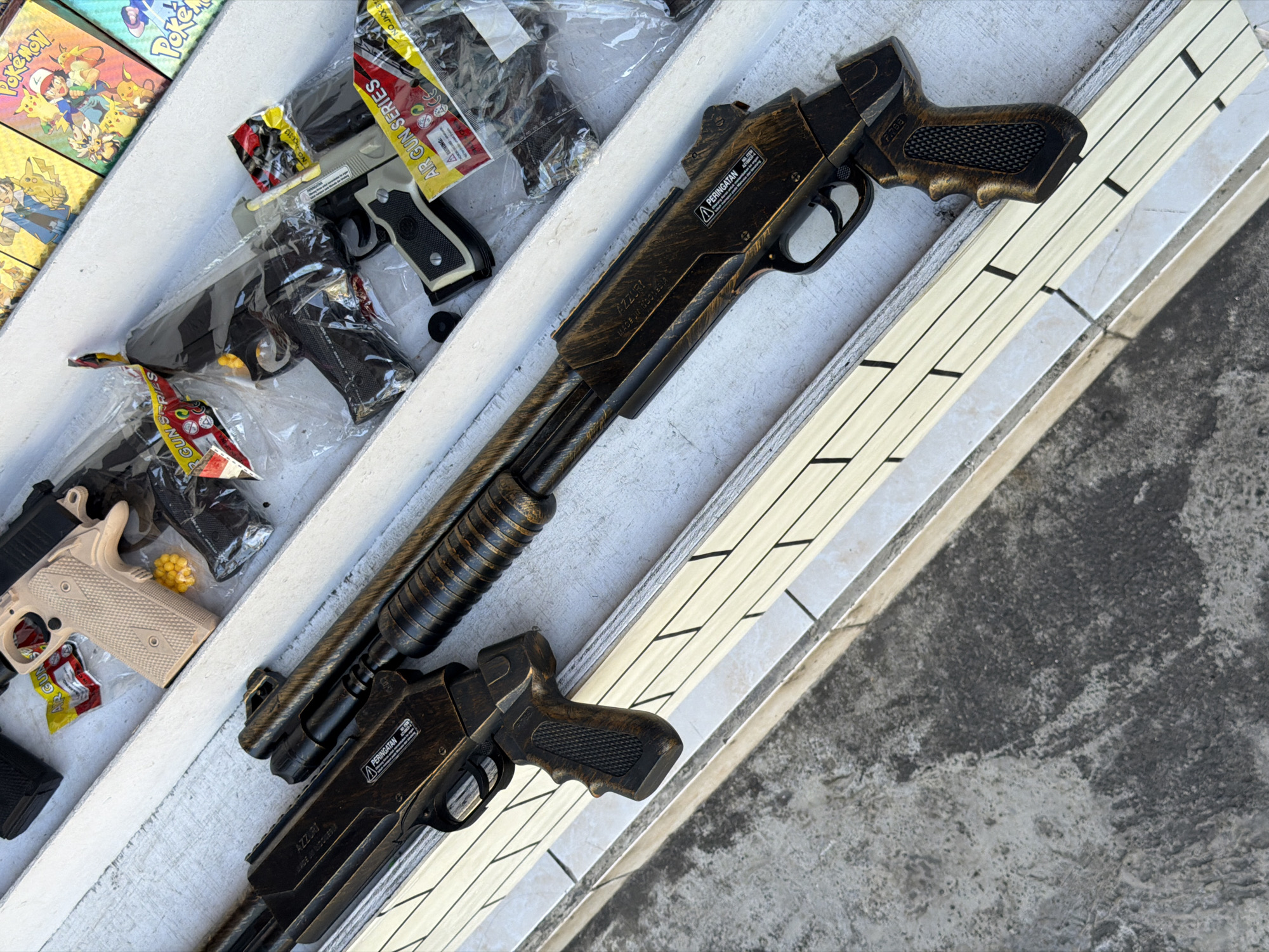 The image displays a collection of toy guns arranged on a surface with white and striped sections. There are several plastic-wrapped toy pistols and a pump-action shotgun-style toy gun. The toy shotgun is mostly dark with gold accents. Some Pokémon-themed items are visible in the upper left corner. Labels such as "AZZURI" and "PERINGATAN" are visible on the toy shotgun. "AIR GUN SERIES" is visible on one of the packages. This appears to be a display of toys for sale, in Kuta, Indonesia, based on the "MADE IN INDONESIA" label on the shotgun.