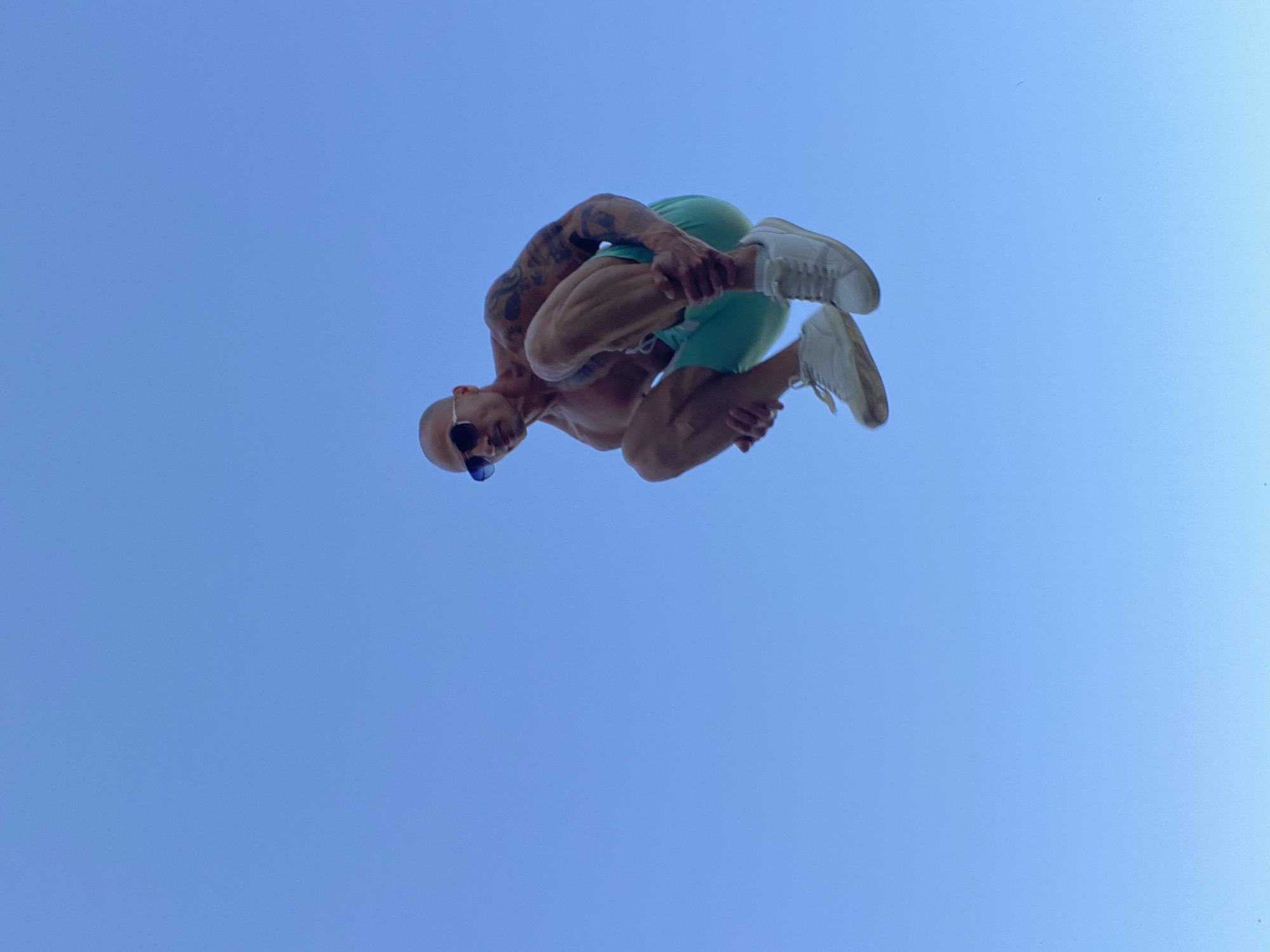 A shirtless, muscular man with tattoos on his arms and sunglasses is captured mid-air, appearing to be jumping against a clear blue sky. He is wearing teal shorts and white sneakers. His knees are tucked towards his chest, and he is holding his shins with his hands. The image does not provide any specific information about the location in the city of Pleven, Bulgaria.