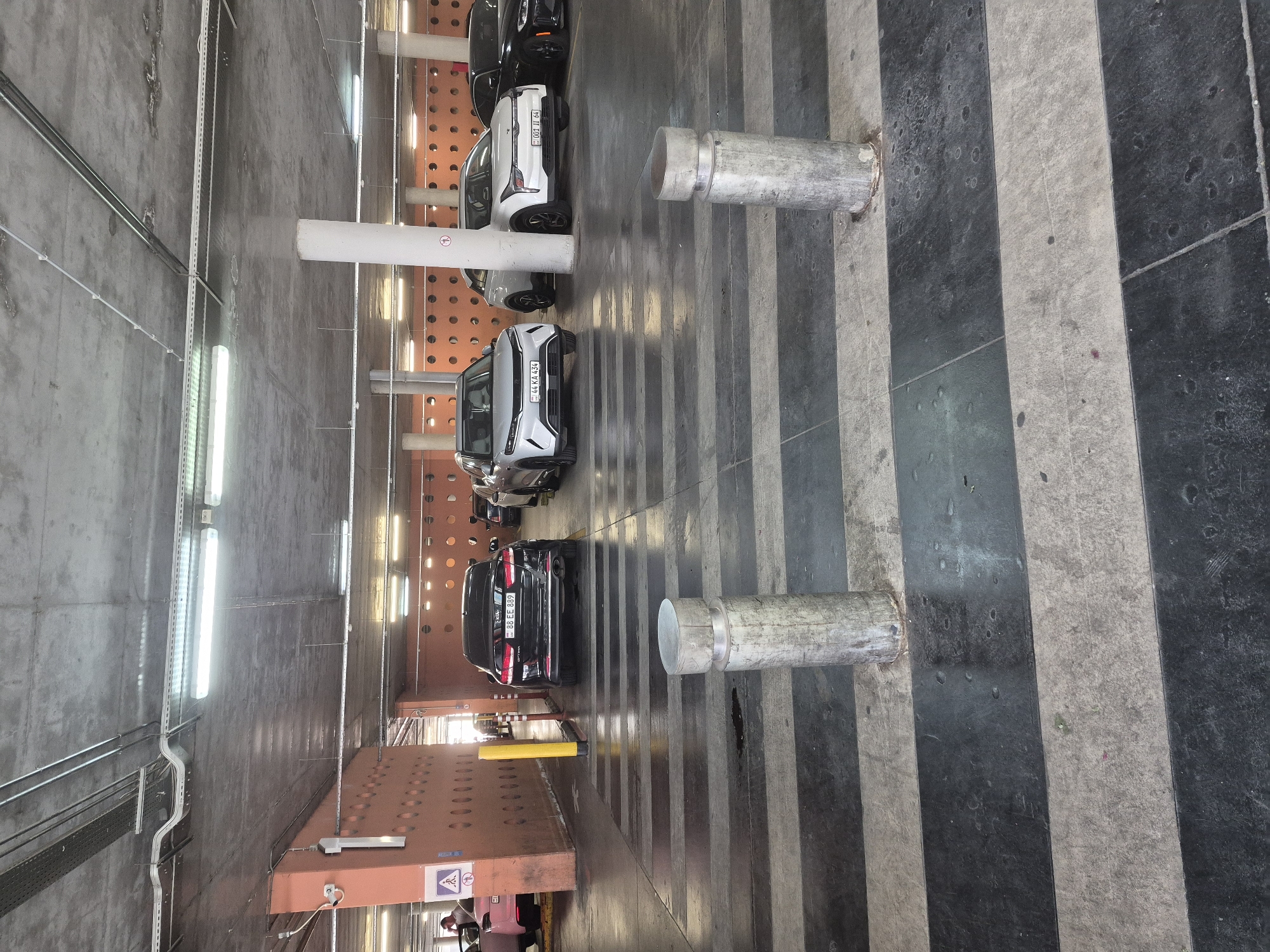 The image depicts an indoor parking garage with several cars. The garage has a concrete ceiling with exposed conduits and fluorescent lighting fixtures. The walls are painted orange and feature a pattern of circular openings. The floor is marked with alternating black and white stripes.
In the foreground, two concrete bollards are visible. Several cars are parked in the garage, including a black KIA with license plate "00 EE 889". To the right of the KIA, a white car with license plate "001 TT 64" is parked, and further to the right, a black car is partially visible. To the left of the KIA, a white car with license plate "44 KA 434" is parked. In the far left of the image, part of a pink vehicle and a person can be seen.
A yellow bollard and a red and white striped bollard are positioned near the orange wall on the left. There is a sign on the orange wall with a symbol of a person walking with a briefcase.
Based on the license plates and the overall context, the location is likely a parking garage in Yerevan, Armenia. The license plates visible (e.g., "00 EE 889", "44 KA 434", "001 TT 64") are consistent with Armenian license plate formats.