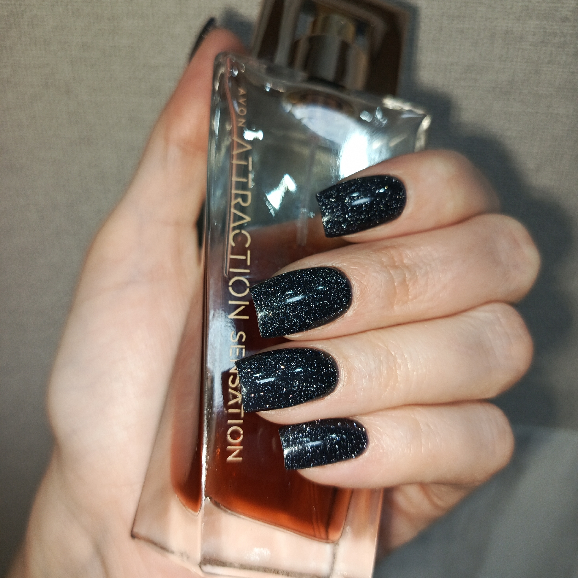 A hand holds a bottle of Avon Attraction Sensation perfume. The background is a neutral color. The location is unknown and cannot be determined from the image.