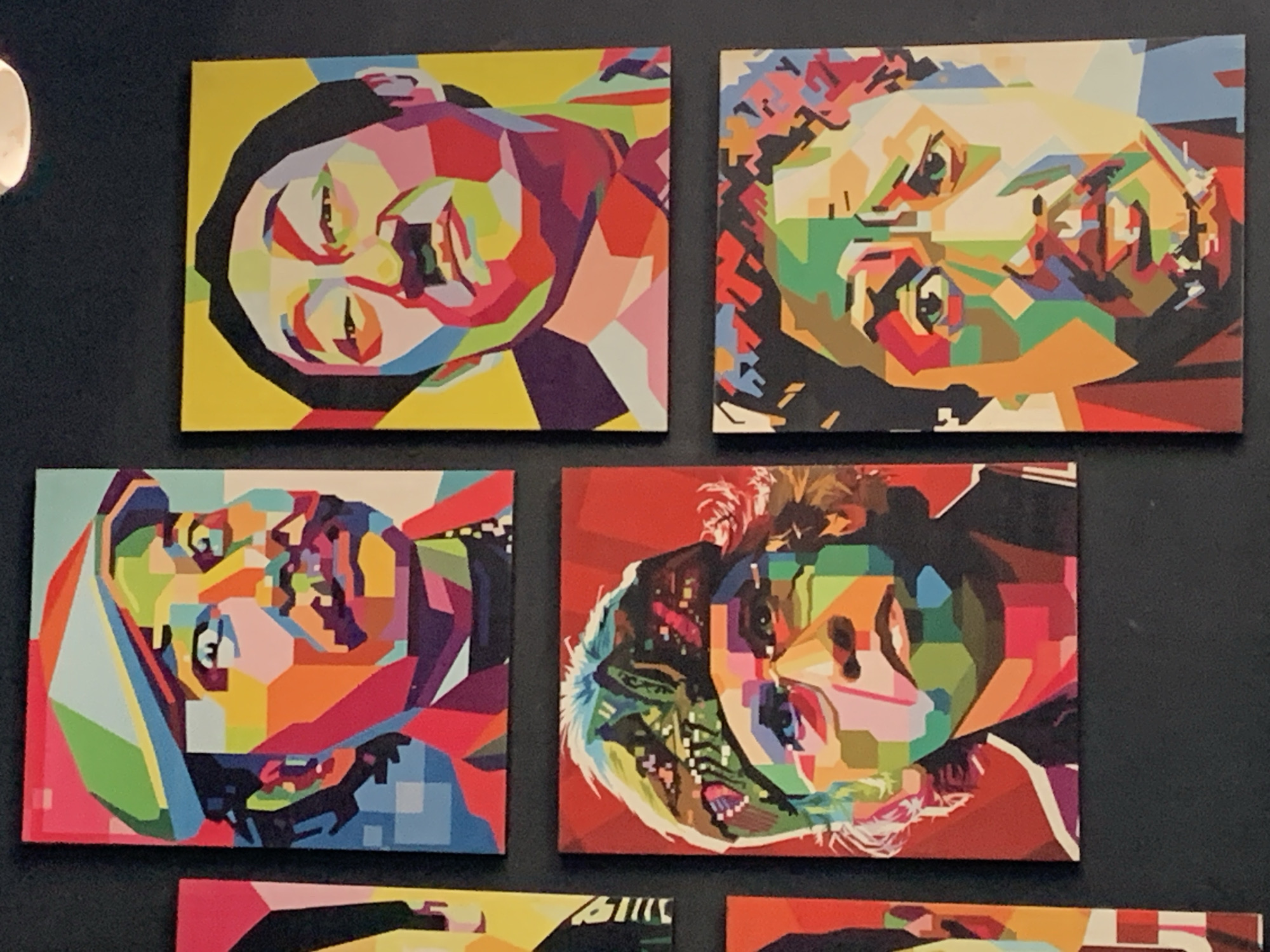 The image displays a collection of five pop art portraits of men, arranged in a grid on a dark wall. The artworks are characterized by bold, geometric shapes and vibrant, contrasting colors. From top-left, moving clockwise, the portraits are:
1. **Top-left:** A portrait of a man wearing a multi-colored cap, with a mustache and goatee. He has a serious expression. The background is a muted teal.
2. **Top-right:** A portrait of a man with dark hair and a mustache, looking slightly downward. The background is bright yellow.
3. **Middle-right:** A portrait of a man with a bandana on his head, looking directly at the viewer. His face is rendered in a mosaic of colors. The background is red.
4. **Bottom-right:** A close-up portrait of a man with a mustache and a beard stubble. His eyes are looking forward. The background is black.
5. **Bottom-left:** Partially visible, this portrait appears to be of a man with fair skin and blonde hair, facing left. The background is red.
The overall presentation suggests an art exhibition or a decorated wall in a public or private space. The specific location mentioned, Palohlada, Indonesia, is not visually discernible from the image itself; this information would need to be contextual.