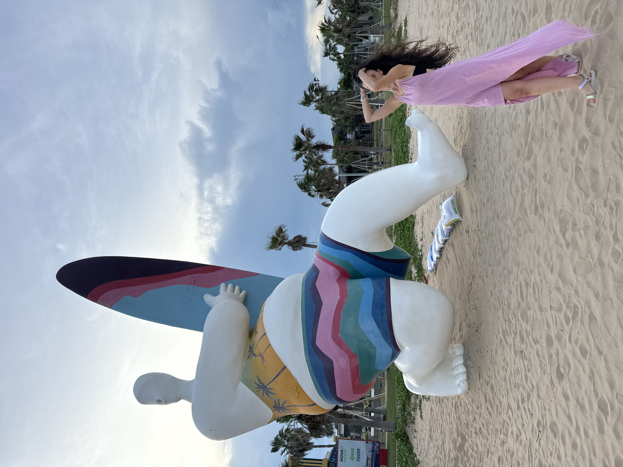A large, white, stylized sculpture of a person with a surfboard is positioned on a sandy beach in Tien Thanh Ward, Vietnam. The sculpture is depicted in a kneeling pose, with colorful striped shorts and a tropical-patterned midriff. To the right of the sculpture, a woman with long, dark hair is standing on the sand, wearing a flowing pink dress. She has her hands raised behind her head and is looking up at the sky. In the background, palm trees and a cloudy sky are visible. A sign for "Nova Beach" and an arrow pointing to "700m" can be seen to the left of the sculpture. There are also several sandbags lined up on the beach near the sculpture's feet.