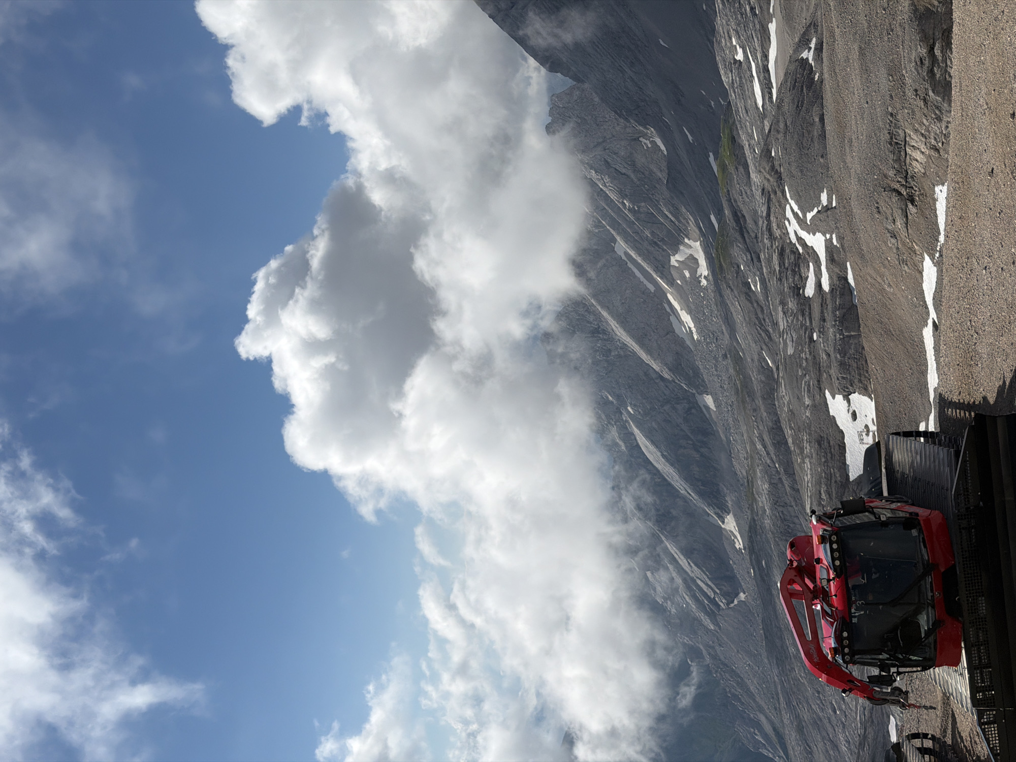 The image depicts a bright, sunny day in the mountainous region of Garmisch-Partenkirchen, Germany. In the foreground, a vibrant red snow groomer is parked on a gravelly surface. The snow groomer has a distinctive arm mechanism extending from its front. The background is dominated by a majestic mountain range, with steep, rocky slopes partially covered in scattered patches of snow. Dramatic white clouds are suspended in a clear blue sky, casting shadows on the rugged terrain below. The overall scene conveys a sense of natural beauty and the ruggedness of alpine environments, with a man-made element in the form of the snow groomer.