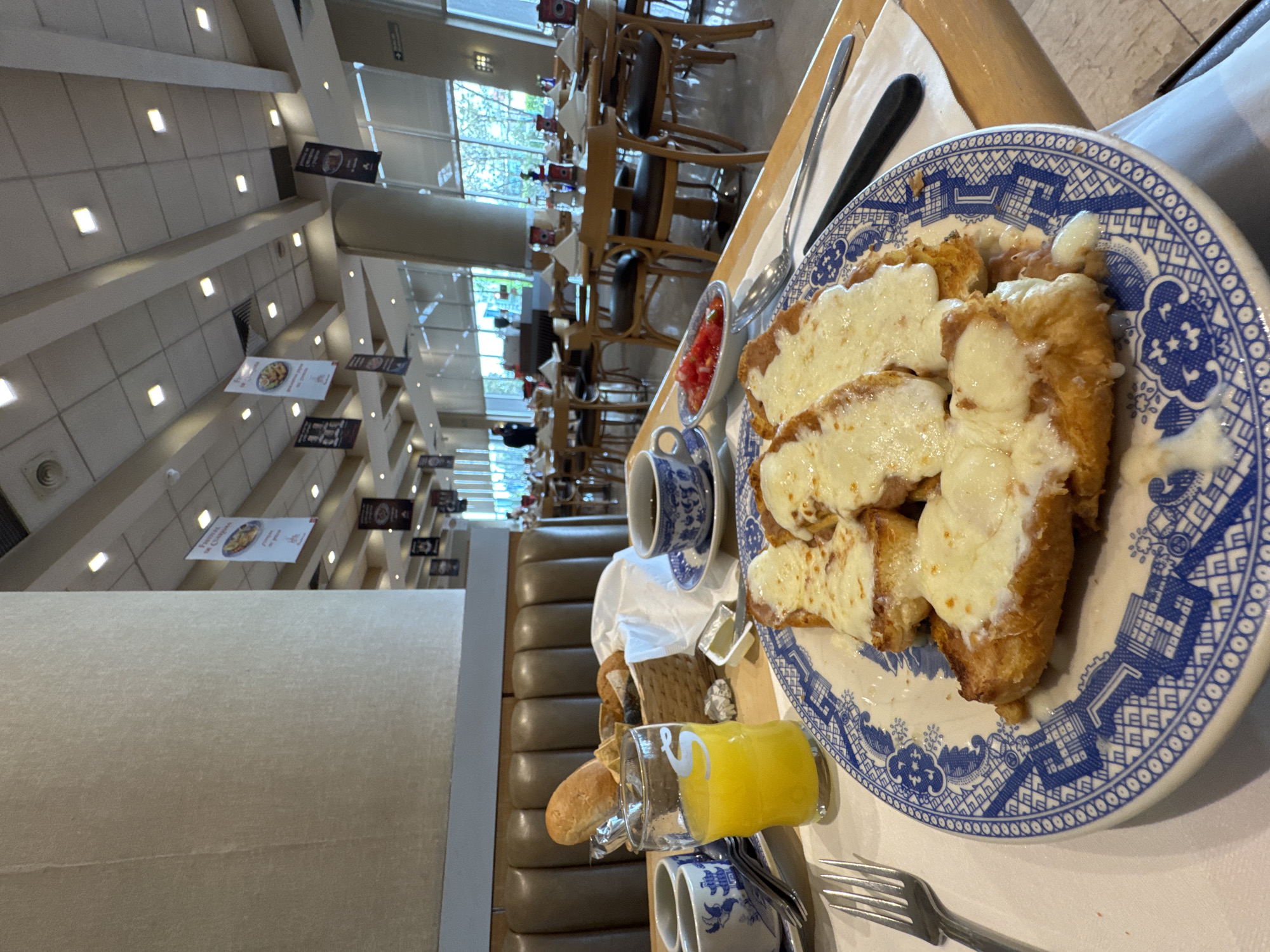 The image depicts a meal at a restaurant, likely in Puebla, Mexico. The scene is a table set for a meal, with the primary focus on a blue and white patterned plate holding a dish of bread slices topped with melted cheese. Beside the plate, there's a small dish containing diced tomatoes, a cup and saucer, a basket of bread, and a glass of orange juice. Silverware, including a fork and spoon, is also visible. The table is situated within a restaurant setting, characterized by long rows of tables and chairs extending into the background, with natural light streaming in from windows. The restaurant's interior includes overhead banners and a long booth seat along the side. Patrons can be seen dining at other tables in the distance.