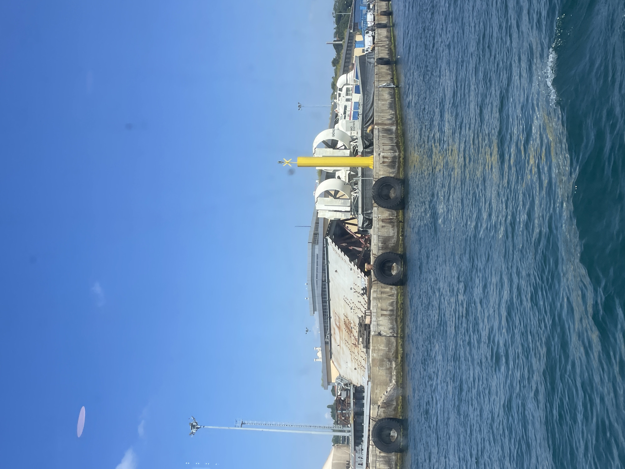 A waterfront scene in Istanbul, Turkey, features a harbor with a large hovercraft docked at a pier. The hovercraft, identified by the markings "GH2142" and "IMO 9327891", is a white and blue vessel with large fans on top. It is positioned next to a yellow marker pole. The pier itself is made of concrete and lined with large black tires acting as fenders. Behind the pier, a damaged or under-construction ramp structure is visible, along with various industrial buildings and some vegetation. The sky is clear blue with a few wispy clouds. The foreground is dominated by the deep blue water of the harbor, with gentle ripples on its surface.