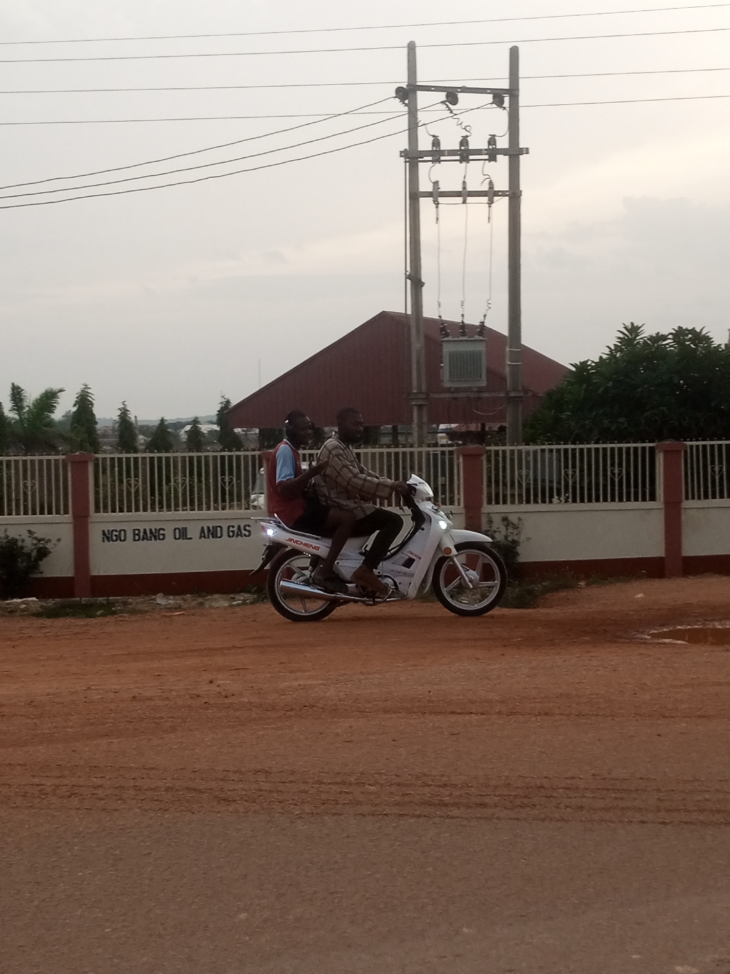 Two individuals are riding on a white motorcycle traveling from left to right on a dirt road. The person in front is driving, wearing a plaid long-sleeved shirt and dark pants. The person behind is wearing a blue short-sleeved shirt and a maroon vest, and is looking back towards the viewer. The motorcycle has the brand "JINCHENG" visible on its side. In the background, there is a white fence with a sign that reads "NGO BANG OIL AND GAS". Behind the fence, a large building with a dark red roof is visible, as well as trees and utility poles with electrical wires. The sky is overcast. The scene appears to be in an outdoor, possibly semi-rural or industrial area.