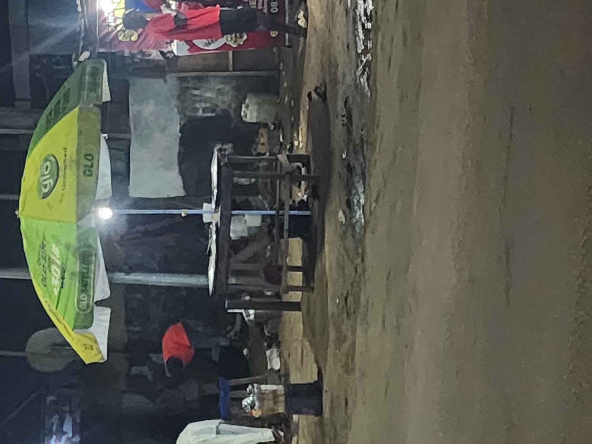 The image depicts a street scene in Obiaruku, Nigeria. The focus is on a simple outdoor eating area with wooden tables and chairs, partially covered by a large, advertising umbrella for the GLO telecommunications company. People are present in the background, though details are obscured by shadow and distance. The ground appears to be a mix of dirt and pavement. The atmosphere seems to be low-light, possibly evening. There is no indication of any interactions occurring in the image.