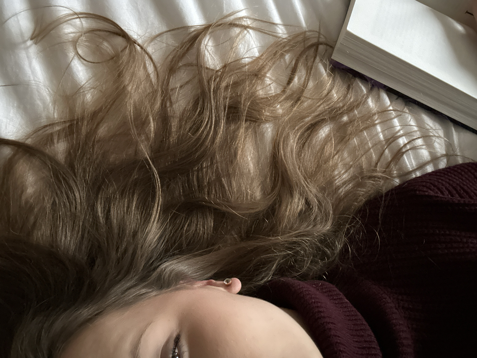 A close-up shot of a person lying on a white sheet, with their head towards the bottom of the frame. Only a portion of their face, including the forehead, eyebrow, and part of an eye, is visible. They are wearing a dark maroon knit sweater. The person's long, light brown wavy hair is spread out across the sheet. A thick, white book with a hint of purple on its side is positioned near the top right corner of the frame. The environment appears to be indoors, possibly a bedroom, based on the bedding and personal items. There is no information to determine this is in Truchtersheim, France.
