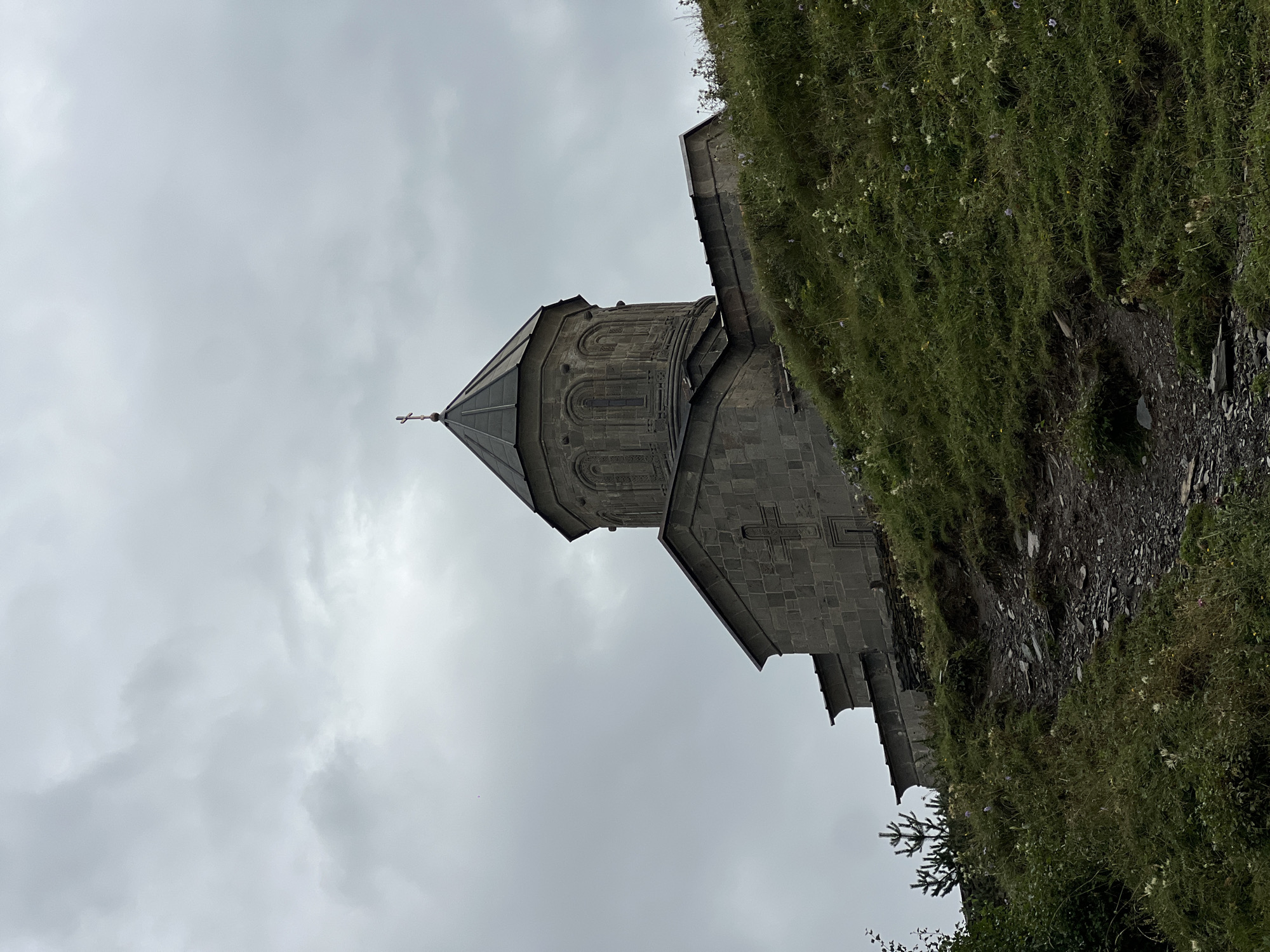The image shows Gergeti Trinity Church, located in Stepantsminda, Georgia, on a cloudy day. The church is constructed of stone and sits on a grassy hill. A path made of rocks and dirt leads up the hill towards the church. The sky is overcast with gray clouds. Green grass and small white wildflowers cover the hillside. A small evergreen tree is visible to the left of the church. The architecture of the church is characterized by a central dome topped with a cross. The walls have stone construction and decorative elements, including a cross carved into the facade.