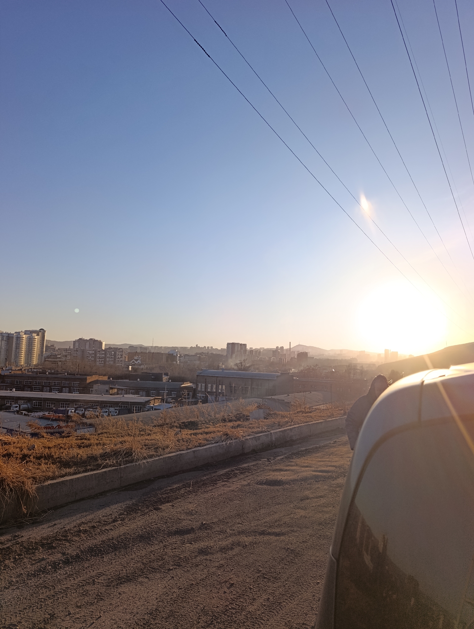 A photo taken at sunset in Krasnoyarsk, Russia shows a view of the city from an elevated position, likely a road. A portion of a car's exterior is in the foreground on the right side of the image. In the mid-ground, there is a low concrete wall, a patch of dry vegetation, and a collection of low-rise industrial or commercial buildings with parked vehicles. Behind these, the cityscape emerges with taller residential and commercial towers visible. A low mountain range is faintly discernible in the background. Electrical lines stretch across the clear, light blue sky, and the setting sun casts a bright glow. The road in the foreground shows tire tracks in the dirt. There are no people visible, apart from the possible reflection of a person on the car's exterior.