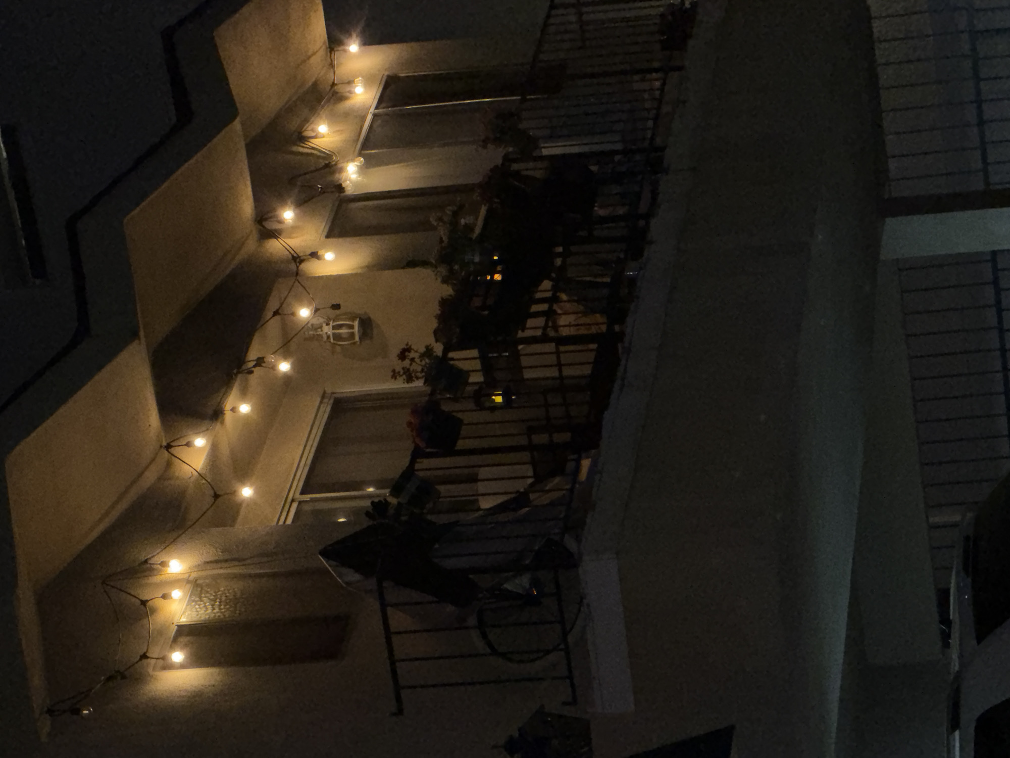 An exterior nighttime shot shows a balcony with string lights illuminated across the top. Below the lights is a railing, and on the balcony, there are plants and what appears to be a grill covered by a tarp. To the left of the balcony, a white lantern is mounted on the wall. Below the balcony, a dark fence is visible, and a portion of a white SUV is in the foreground. The overall scene is dimly lit by the string lights.