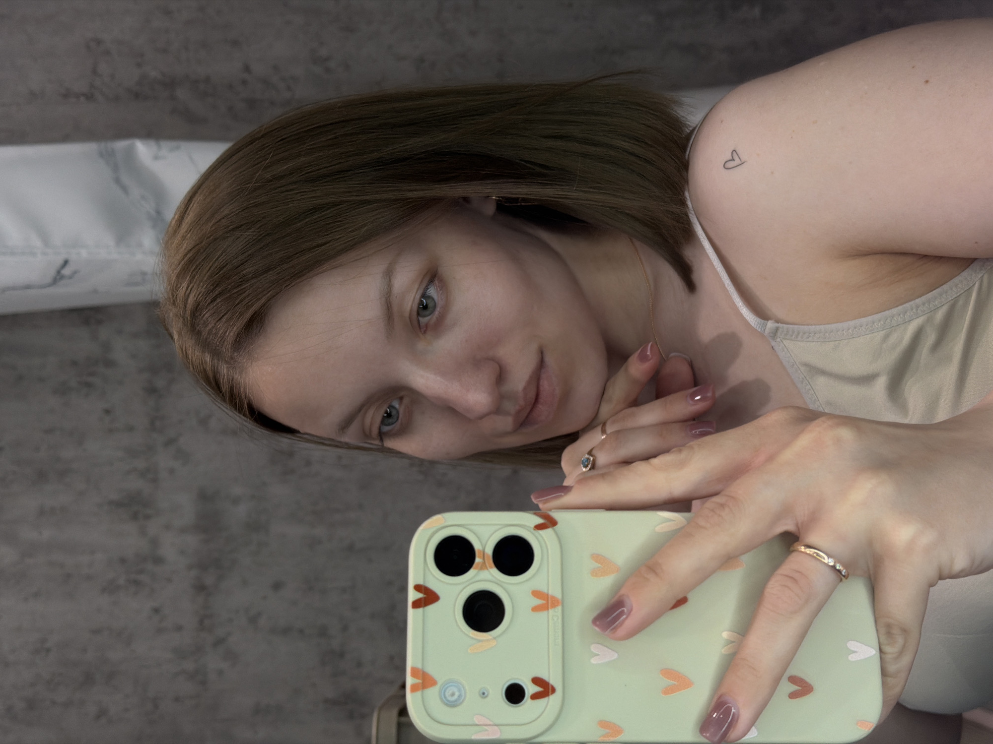 A woman is taking a selfie with her smartphone. She is wearing a beige tank top and has a small heart tattoo on her left shoulder. Her hair is short and brown, and she has light-colored eyes. She is wearing two rings, one on her ring finger and one on her pinky. The smartphone has a light green case with small heart designs. The background is a grey wall, with what appears to be a white radiator above it. The location is Reshetnikovo, Russia.