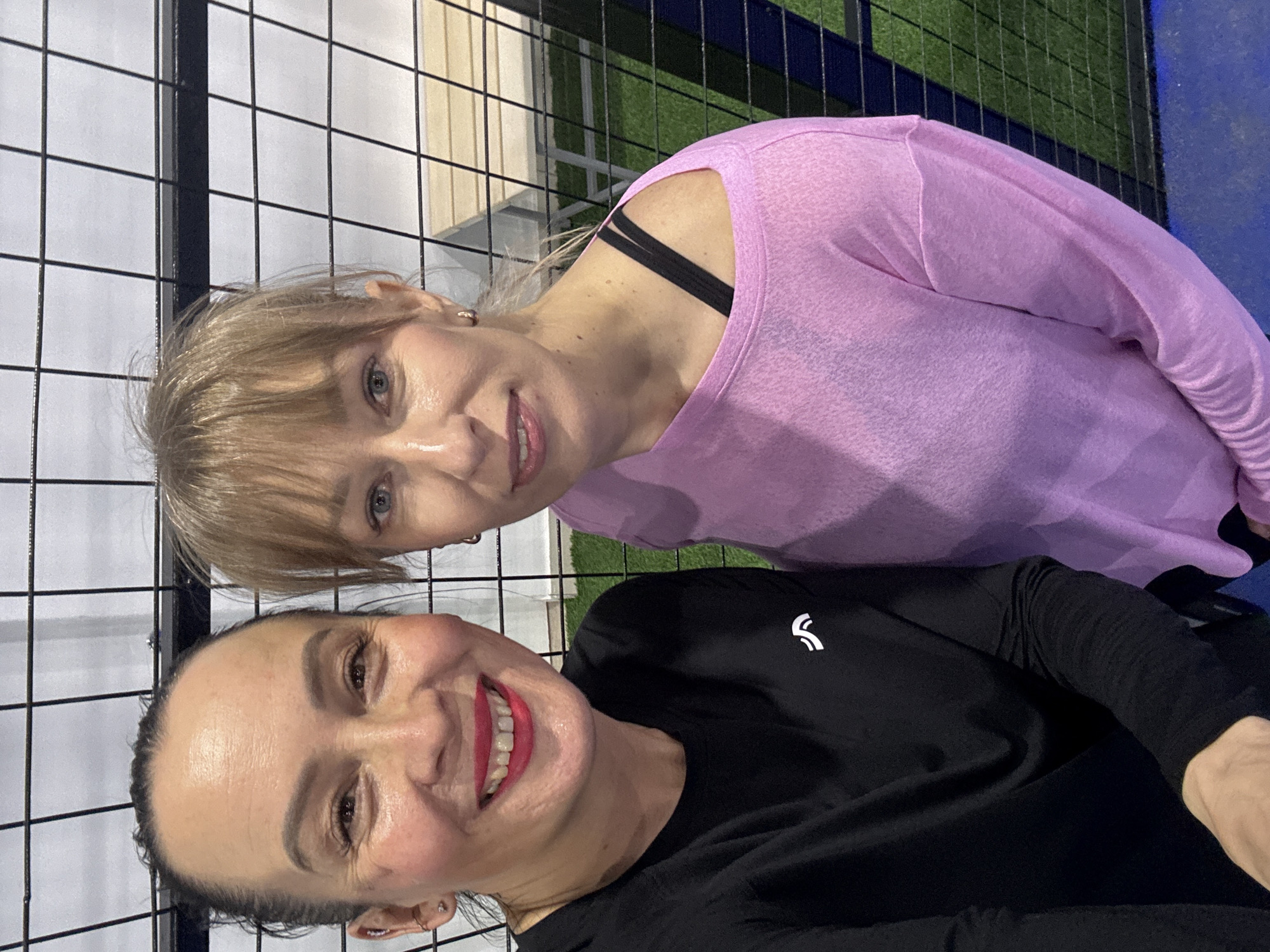 Two women are posing for a selfie in what appears to be an indoor sports facility or Padel court, as suggested by the black metal fence and artificial turf visible in the background.
The woman in the foreground is wearing a black long-sleeved shirt with a small white logo on the chest. She has dark hair pulled back, fair skin, and is wearing bright red lipstick. She is smiling broadly. The woman behind her is wearing a pink shirt that reveals a black bra strap on her shoulder. She has blonde hair with bangs and is also smiling at the camera.
Based on online information, the location is likely Chaikovsky, Russia, as specified in the prompt. The background includes a white wall and a section of artificial green turf, potentially indicating they are inside a Padel court or similar sports venue.