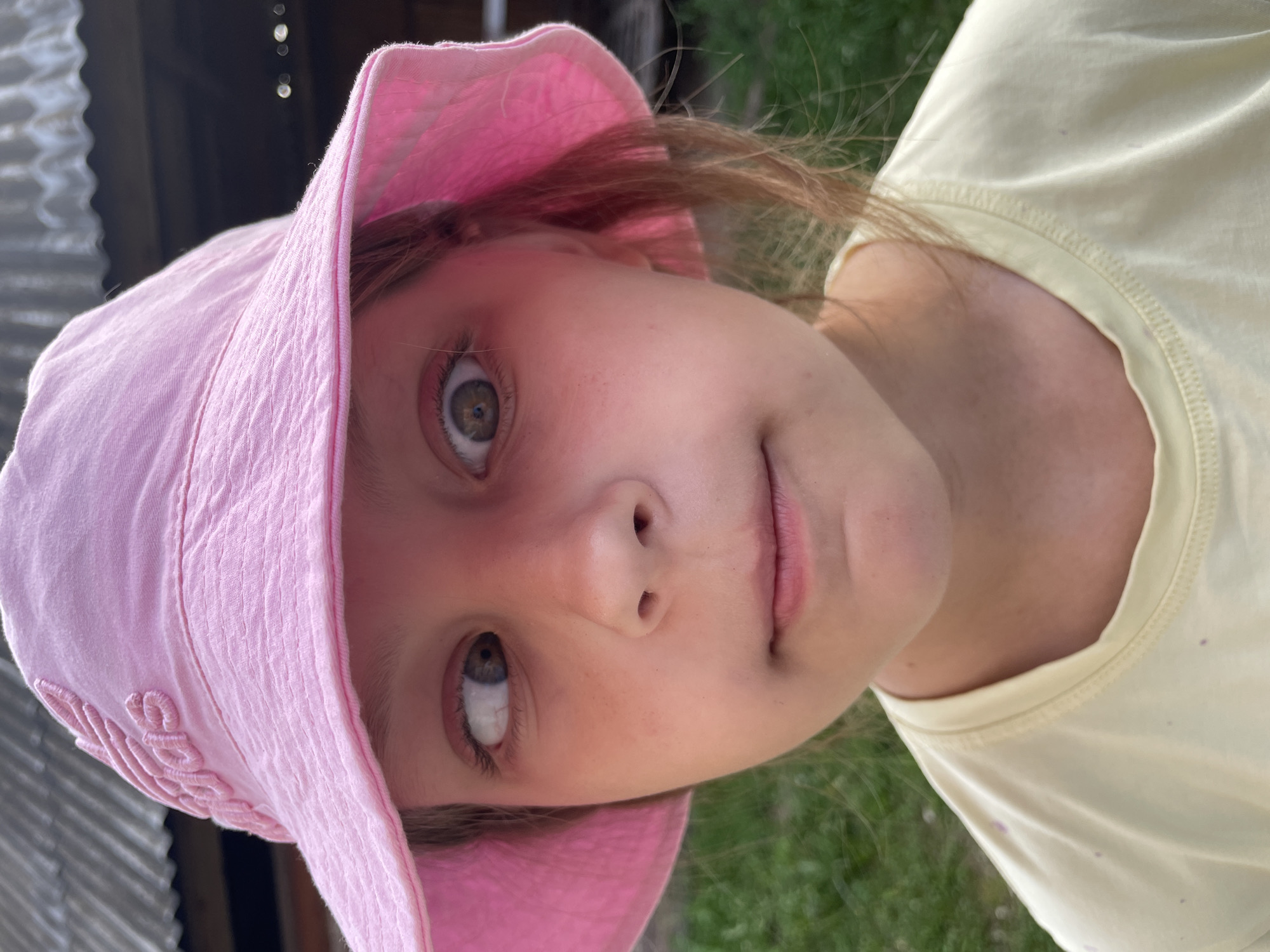 A young girl wearing a pink bucket hat with an embroidered logo and a yellow t-shirt is pictured outdoors. Her eyes are crossed, with one looking to the left and the other looking straight ahead. The background shows blurred green foliage and a corrugated metal structure, suggesting an outdoor setting possibly in a rural or suburban area. The location is not specified within the image data.