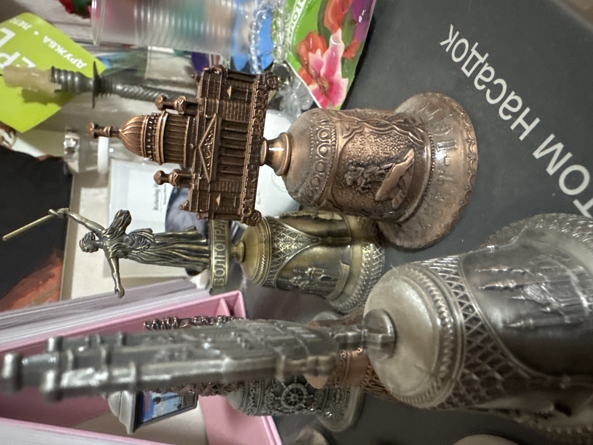 The image shows a collection of decorative bells displayed on a dark surface, possibly a shelf or table. In the foreground, a large, silver-colored bell is prominently featured, adorned with intricate architectural details resembling a cityscape. To its right, a smaller, copper-colored bell is visible, topped with a detailed model of a building with a dome and columns. This bell also features relief carvings of figures and architectural elements around its body.
Behind these bells, several other bells of varying sizes and metallic finishes (bronze and silver) are arranged. One bronze bell in the mid-ground has a figure of a winged woman holding a sword, with text below it that appears to be Russian. Another silver bell to the left has a lighthouse-like structure and a ship's wheel motif. The background is cluttered with various objects, including what looks like books or binders with pink covers, a candlestick with a lit candle, a glass or plastic container, and a green package with floral imagery. The overall impression is of a display of souvenir or decorative bells, likely collected or arranged in a personal space. The exact location is not determinable from the visual information provided.