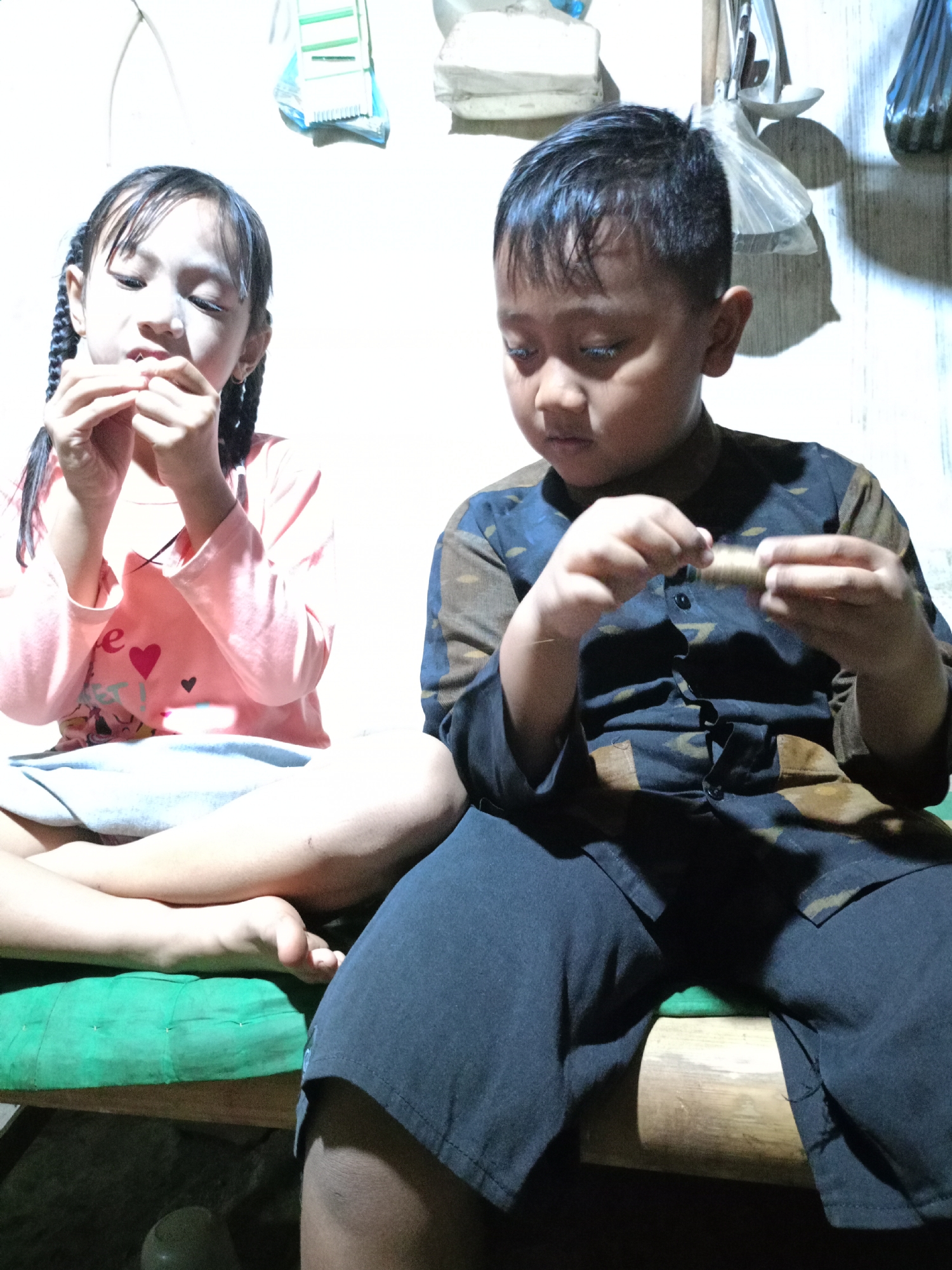 In Jelbuk, Indonesia, two young children, a girl and a boy, sit side-by-side indoors on a green cushioned bench. The girl, on the left, has her hair in braids and is wearing a pink long-sleeved shirt with cartoon designs and a light blue skirt. She is holding a small object to her mouth, seemingly engaged in eating or playing with it. The boy, on the right, is wearing a dark shirt with a patterned design and dark pants. He is holding a small, cylindrical object in his hands, examining it. The background shows a lightly colored wall with some objects hanging from it, partially obscured by shadows. The lighting appears to be artificial, casting shadows on the wall and emphasizing the children's activities.