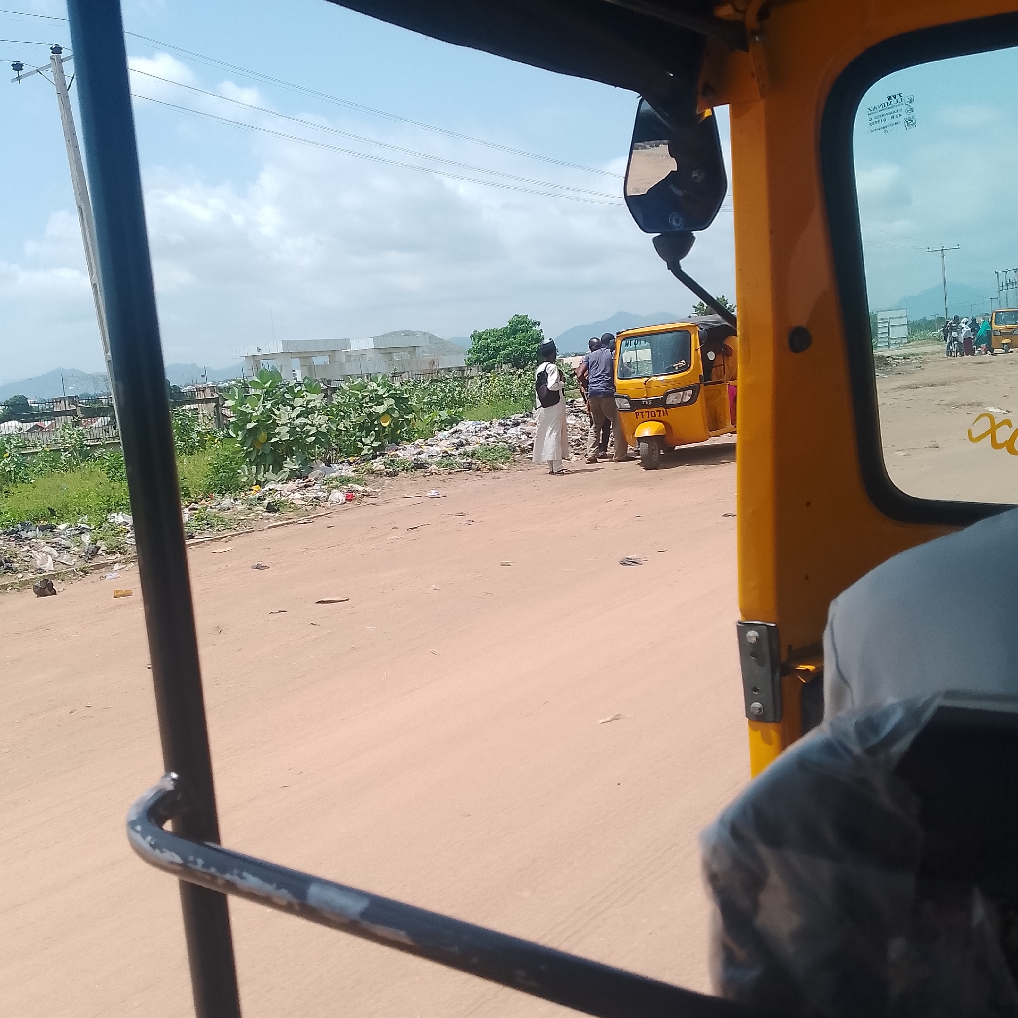 The image depicts an outdoor scene in Jalingo, Nigeria, likely during the daytime given the bright sky and shadows. The perspective is from inside a yellow vehicle, possibly an auto-rickshaw or tuk-tuk, looking outwards.
In the mid-ground, a yellow auto-rickshaw is parked on a dirt road. Three people are standing near it. One person, wearing a white dress and a headscarf, stands with their back to the camera. Two other individuals, both men, are facing away from the camera, appearing to engage in conversation or wait for the auto-rickshaw.
The scene is characterized by a significant amount of scattered trash and debris along the side of the road, suggesting an area with poor waste management. Lush green vegetation is interspersed with the trash, and in the background, there are low hills or mountains under a cloudy sky. Construction or unfinished buildings are also visible in the distance, along with utility poles and power lines. Another auto-rickshaw is visible further down the road, along with other people and a motorcycle.
The overall impression is of a typical street scene in a developing urban or semi-urban area. There are no strong indicators of specific emotions, but the environment appears somewhat neglected due to the litter.
Visible text includes "TVS" on the front of the auto-rickshaw, a license plate that appears to read "PT 707H", and some text on the side window of the vehicle from which the photo is taken, which is mirrored and difficult to fully decipher but appears to be vehicle information or branding. Yellow cursive-like markings are also visible on the side of the vehicle in the foreground.