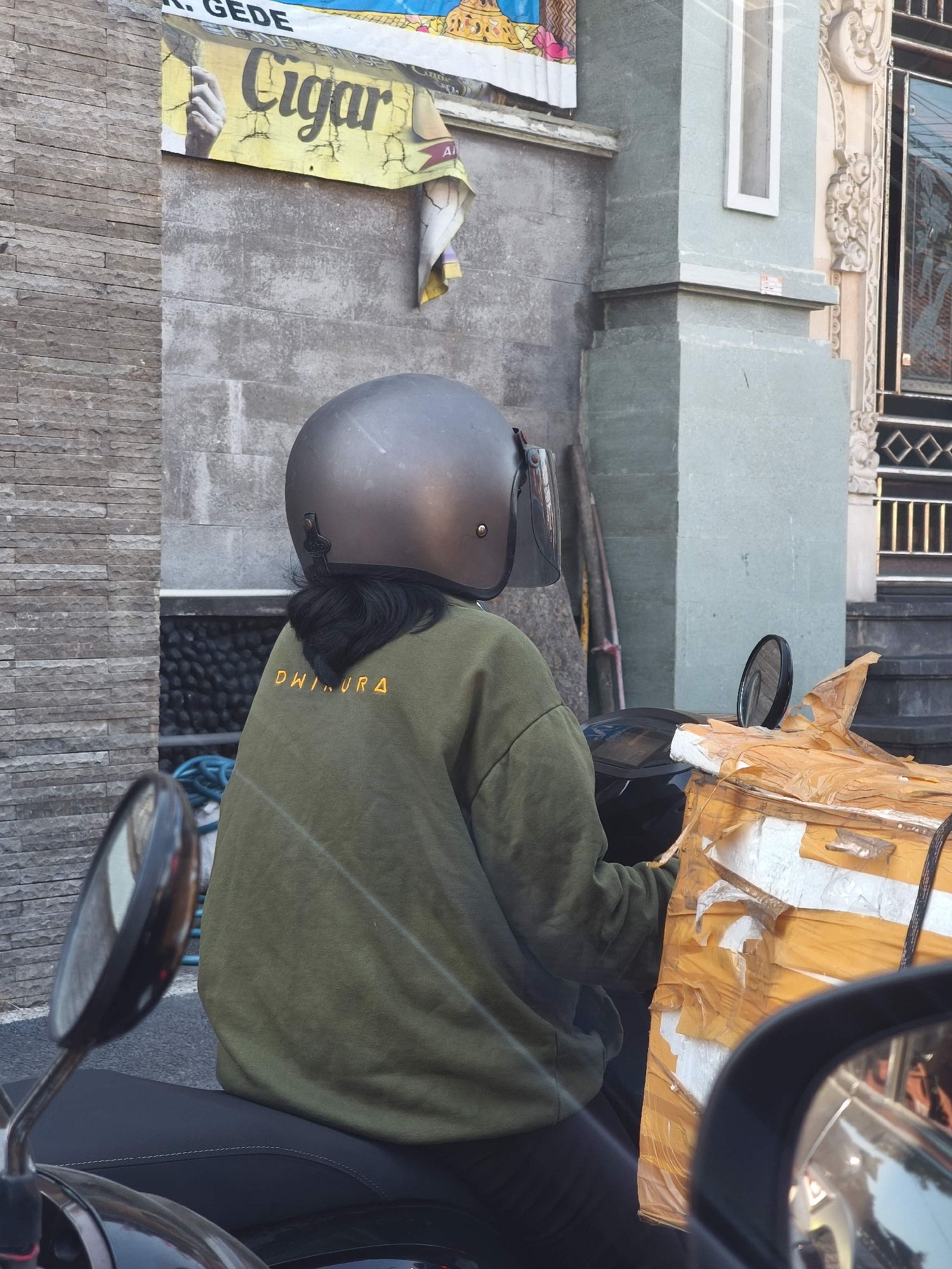 An individual wearing a dark grey helmet and an olive green sweatshirt with "DHIURA" written in yellow lettering on the back is seated on a black motorcycle. The motorcycle is carrying a large, yellow-wrapped package secured with tape. The scene appears to be on a street in North Kuta, Indonesia, with a stone wall and a building in the background. A sign with the word "Cigar" is visible on the wall. A mirror of the motorcycle is in the foreground, reflecting the environment.