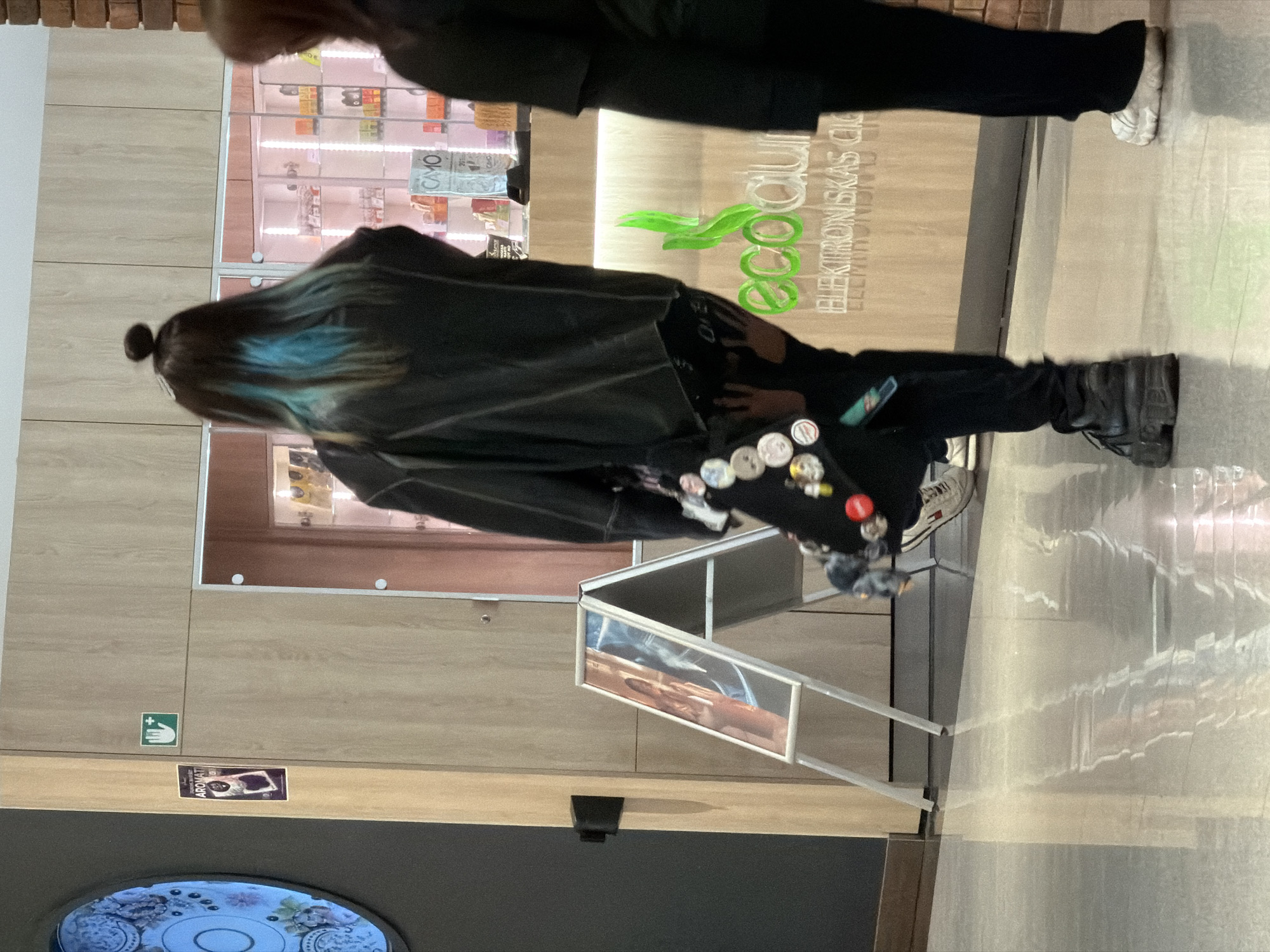 A medium, long shot taken indoors shows a person with dyed blue hair and a bun, a woman wearing black, and a store called "Ecocitrus elektroniskas cigaretes." The person with blue hair is wearing a black jacket, pants adorned with pins, and black boots. They appear to be either crouching or kneeling near a silver A-frame sign. The store features a brightly lit display of products behind glass. The woman wearing black is partially visible at the top of the frame, walking away from the camera. The location is likely a shopping center in Riga, Latvia, based on the Latvian text on the store signage. The overall impression is of a casual scene in a commercial setting.