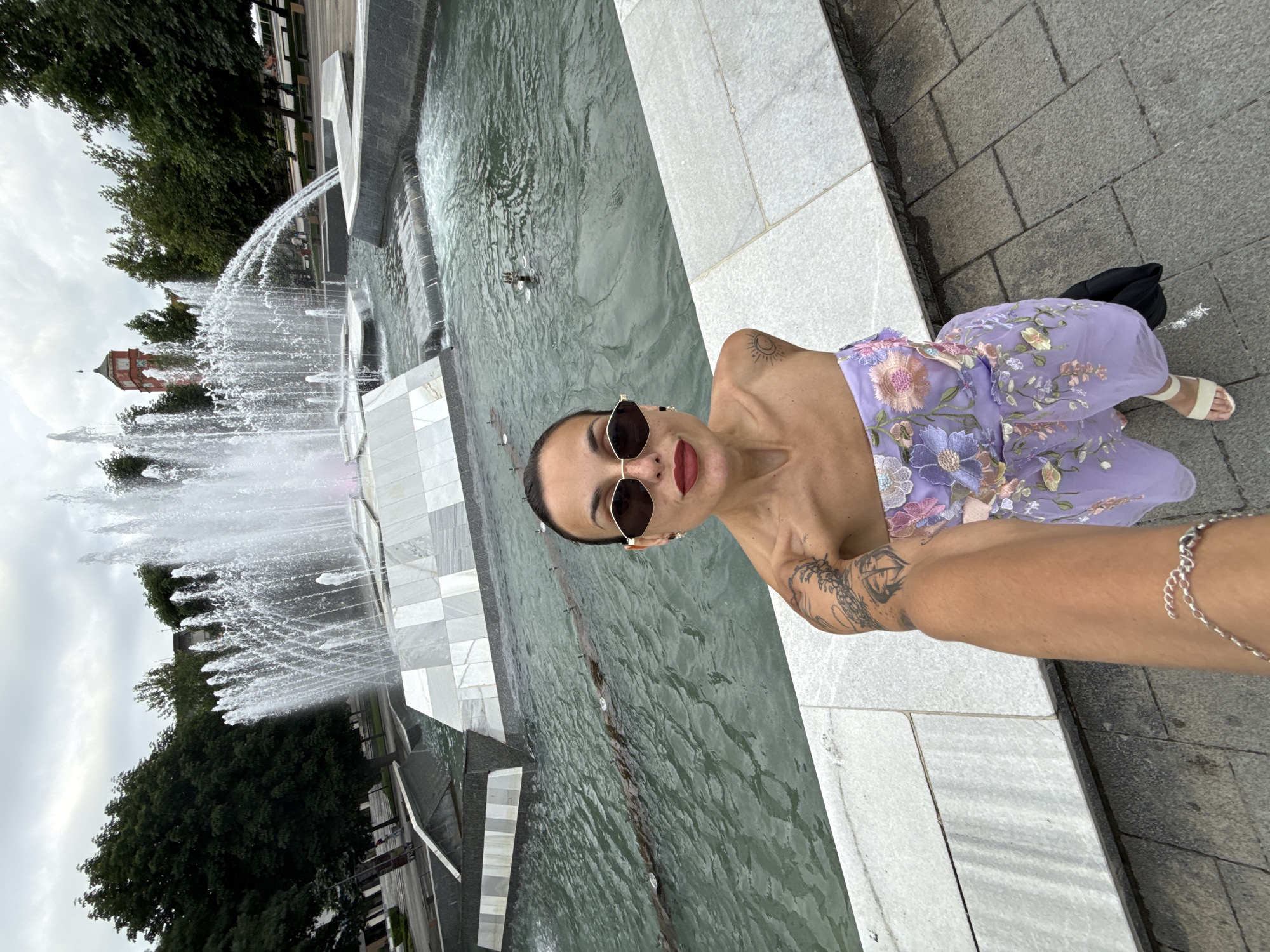 This is a selfie taken by a woman in front of a large, multi-tiered fountain. The fountain has multiple jets of water spraying upwards and outwards, creating a misty effect. The fountain is surrounded by marble and stone structures, and the water is a greenish hue. In the background, there are trees and a red building with a clock tower, which is characteristic of Pleven, Bulgaria. The woman is wearing a strapless lavender dress with floral embroidery and white sandals. She has dark hair pulled back, dark sunglasses, and red lipstick. Her left arm, which is holding the camera, is visible in the foreground, showing tattoos. The sky is overcast.