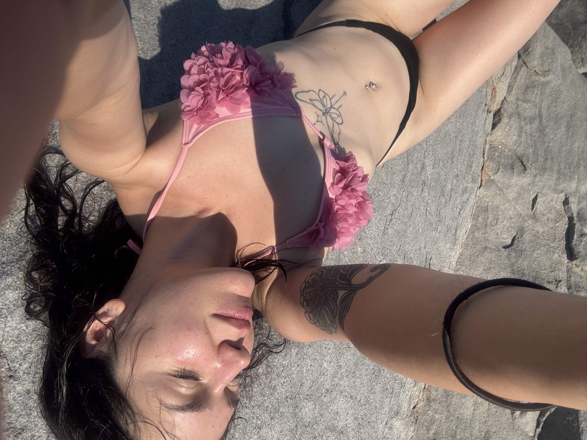 A young woman is lying on her back on a rocky surface, wearing a pink bikini and black bottoms. Her head is tilted back, eyes closed, and her lips are slightly parted. She has a belly button piercing and a tattoo on her side. Her arm is bent, resting above her head, and she has a black band around her upper arm, as well as a tattoo on her forearm. The rocky ground appears to be near the sea, possibly a beach or a breakwater, consistent with the coastal environment of Chernomorets, Bulgaria. The lighting suggests it is daytime, with sunlight casting shadows.