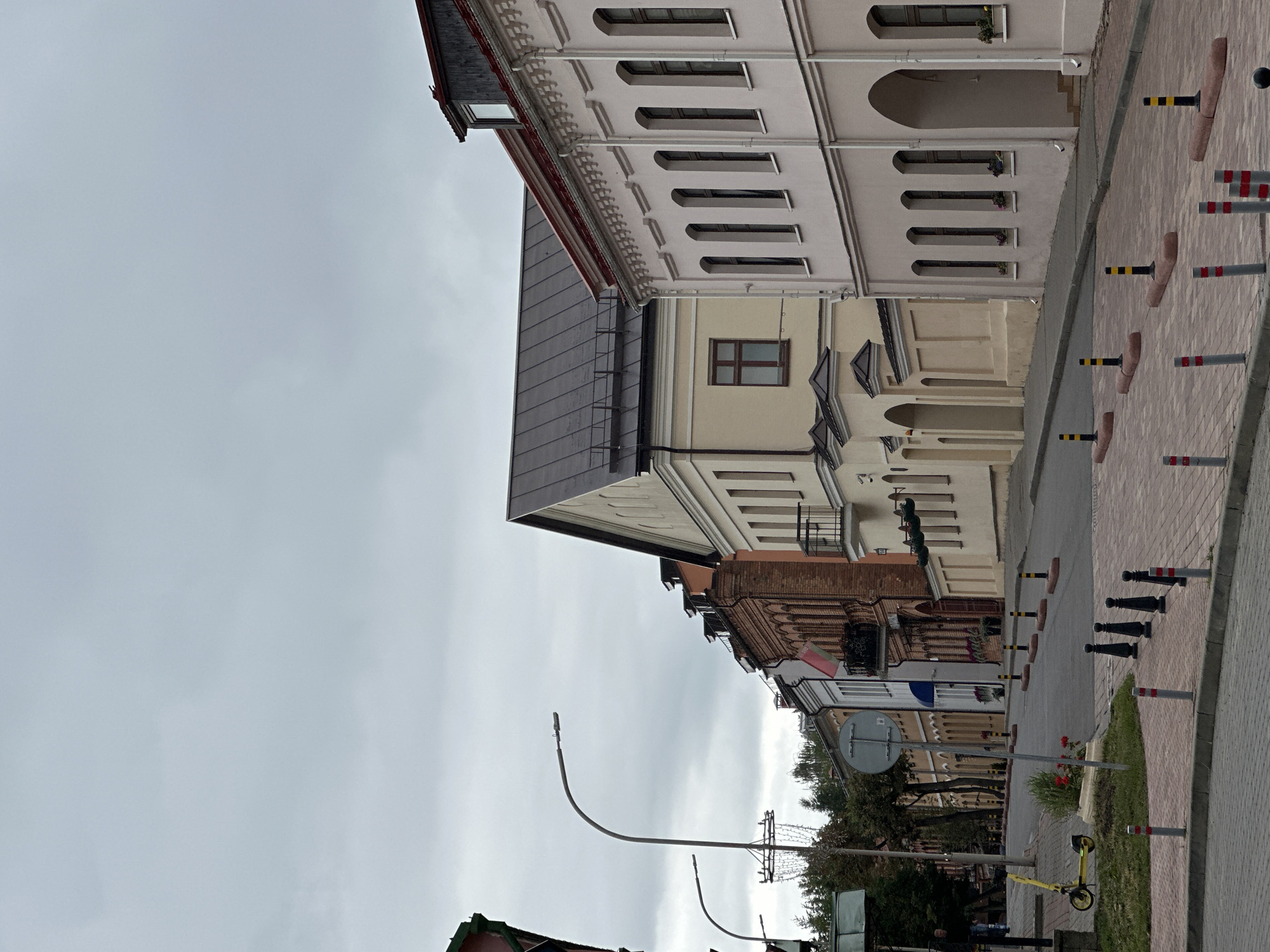 The image depicts a street scene in Minsk, Belarus, under a cloudy sky. The street is paved with red brick, and the sidewalk is also brick. Several bollards, some black and yellow, and others red and white, line the street and sidewalk. A yellow electric scooter is parked on the grass near the sidewalk.
The street is lined with buildings of various architectural styles and colors. The closest building on the right is a light-colored, multi-story structure with arched windows and ornate trim. To its left is a cream-colored building with a dark roof, featuring a visible window. Further down the street, there are buildings with red brick facades, some with balconies, and others with arched doorways. One building displays a white and red flag. Trees are visible along the street, and there are streetlights and utility poles. The overall impression is of an old, well-maintained street in an urban area.