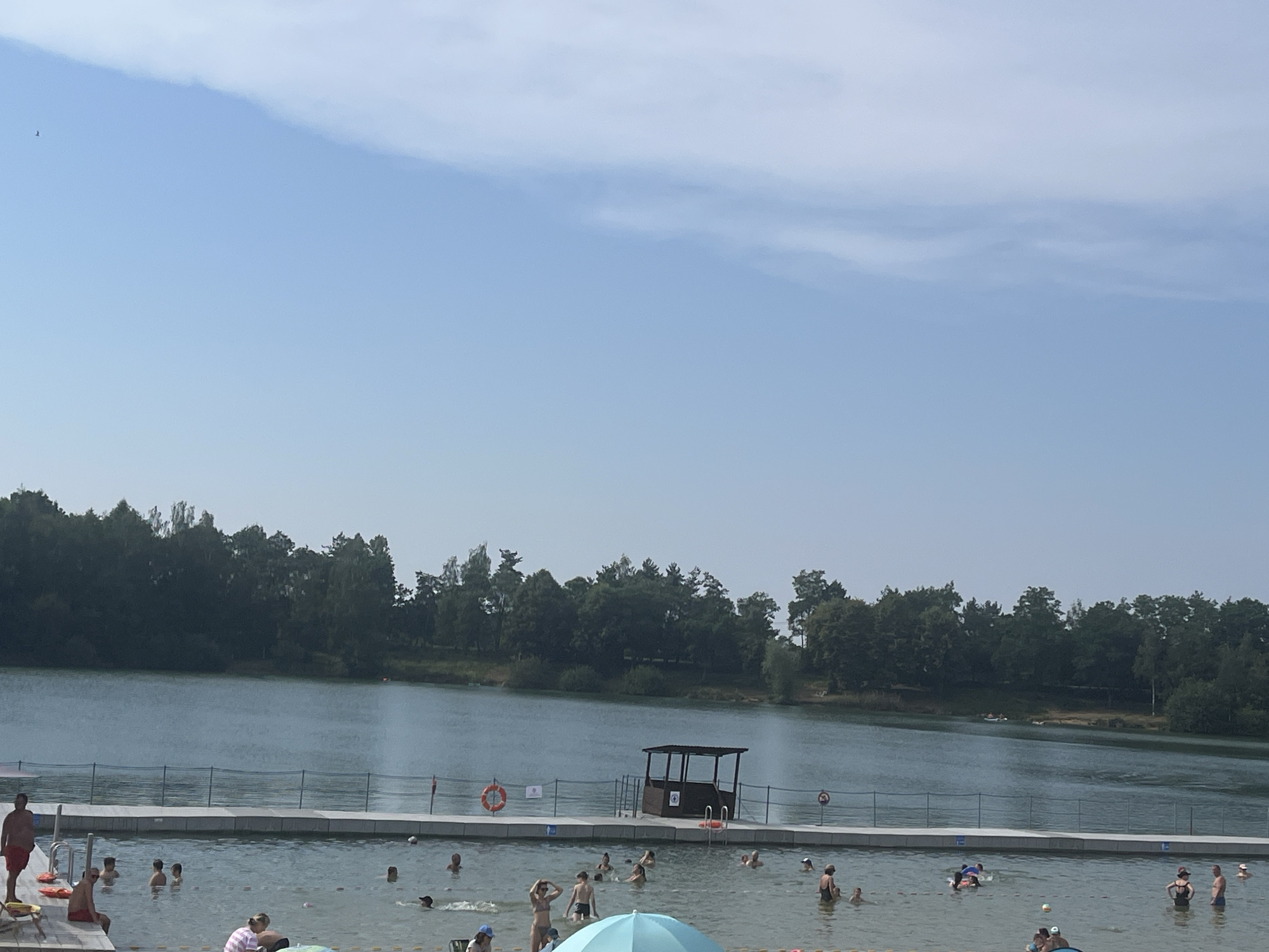 The image shows an outdoor swimming area with a lake in the background and a man-made pool in the foreground. The pool is bordered by a white fence and a wooden deck. Several people are visible in the pool, some swimming and others relaxing. A lifeguard tower is situated on the deck overlooking the pool. The lake is surrounded by a dense line of trees. The sky is clear and blue with a few wispy clouds. The overall scene depicts a recreational area on a sunny day.