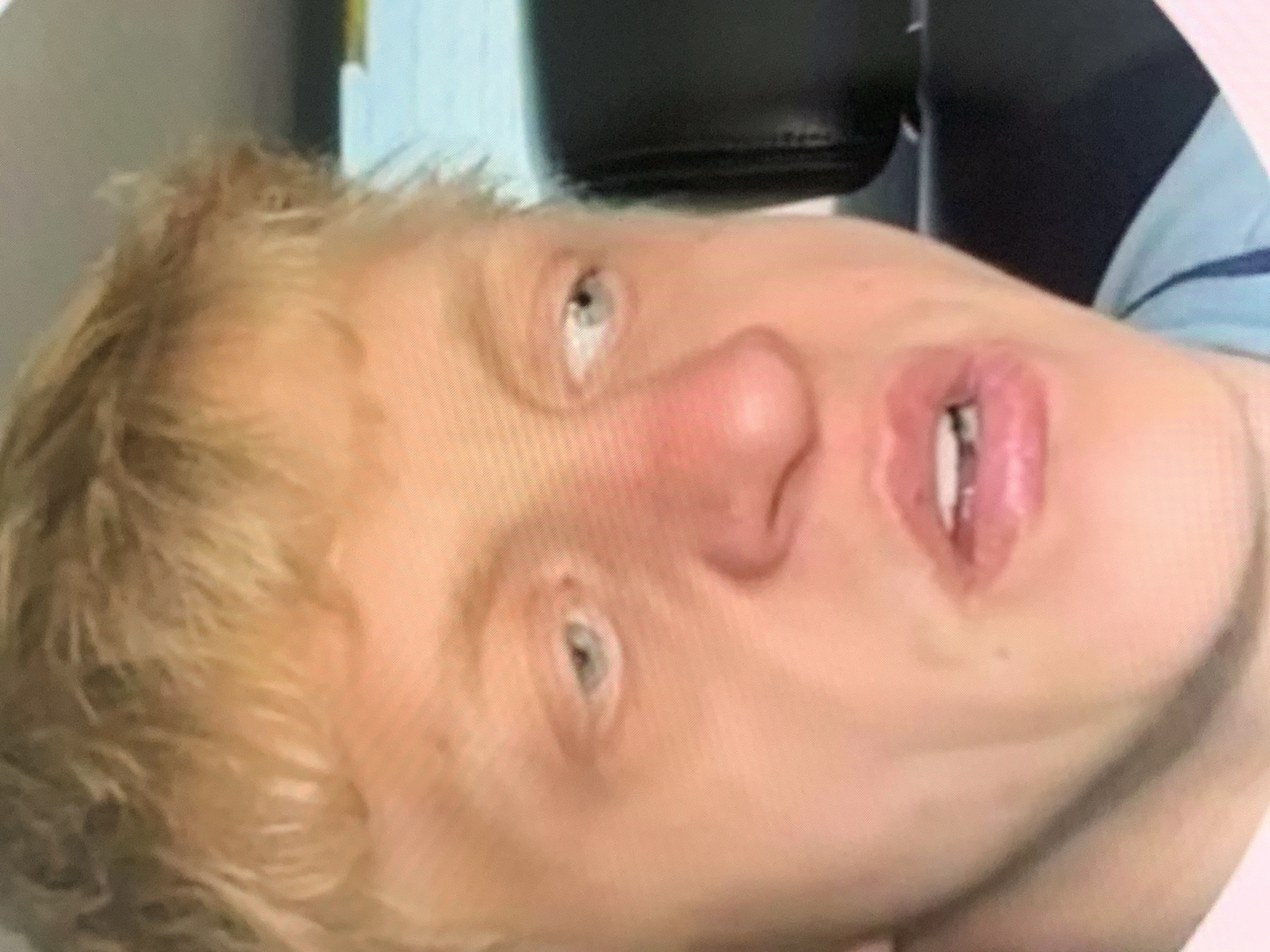 The image is a close-up, low-resolution video still of a young Caucasian male with blond hair. He has light-colored eyes and a reddish nose. His mouth is slightly open, revealing his teeth. The background is blurry and appears to be an indoor setting, possibly a car interior, with a dark headrest visible on the right. There is no identifiable information within the image that places it in the city of Stavropol, Russia.