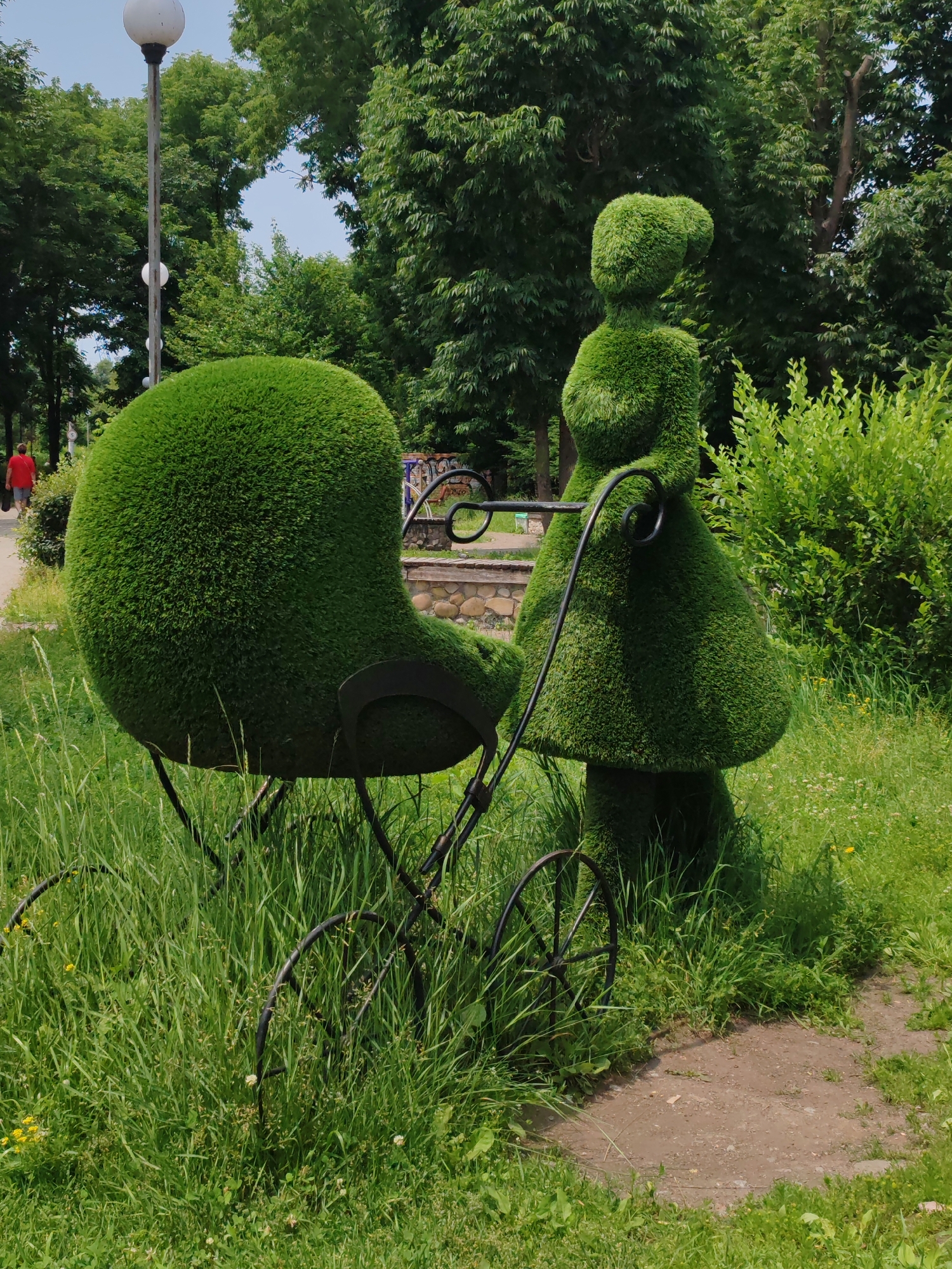 The image depicts a topiary sculpture of a woman pushing a baby carriage in a park setting. The sculptures are made of meticulously trimmed green bushes, forming distinct shapes of a woman wearing a dress and bonnet, and a rounded bassinet-style stroller with black metal accents and wheels. The scene is situated on a green lawn with tall grass and some wildflowers. In the background, there are numerous lush green trees, a stone wall, and a lamppost. A person in a red shirt is walking away from the camera on a path in the distance. The overall environment suggests a bright, sunny day in a well-maintained urban park. While the image's specific location within Vladivostok, Russia, is not discernible from the visual elements alone, the presence of a park with sculptures and trees is consistent with public spaces in the city.