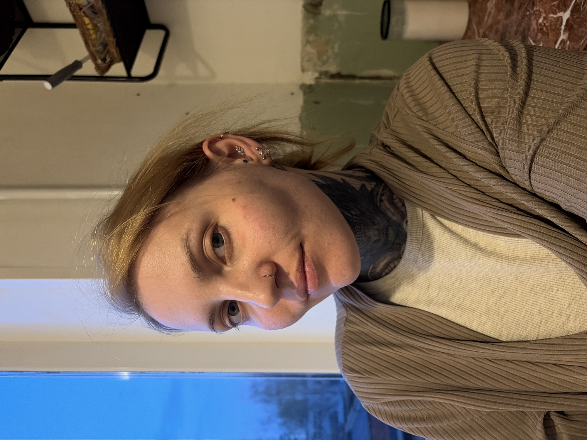 A medium shot depicts a woman with light blonde hair, fair skin, and blue eyes. She has a nose ring and multiple piercings in her right ear, including a hoop, a leaf-shaped design, and several studs. A large, dark tattoo is visible on her neck and chest. She wears a light gray t-shirt and a brown ribbed cardigan.
The background is partially blurred, but appears to include a window with a blue hue at the bottom, suggesting twilight or a blue-toned light source. Above the window, the wall is painted white. To the left, there is a black metal shelf with an object in it. To the right, a wall with green and beige patches is visible. A white cylindrical object rests on a surface that appears to be brown marble. There is no indication of the location being Chaikovsky, Russia.