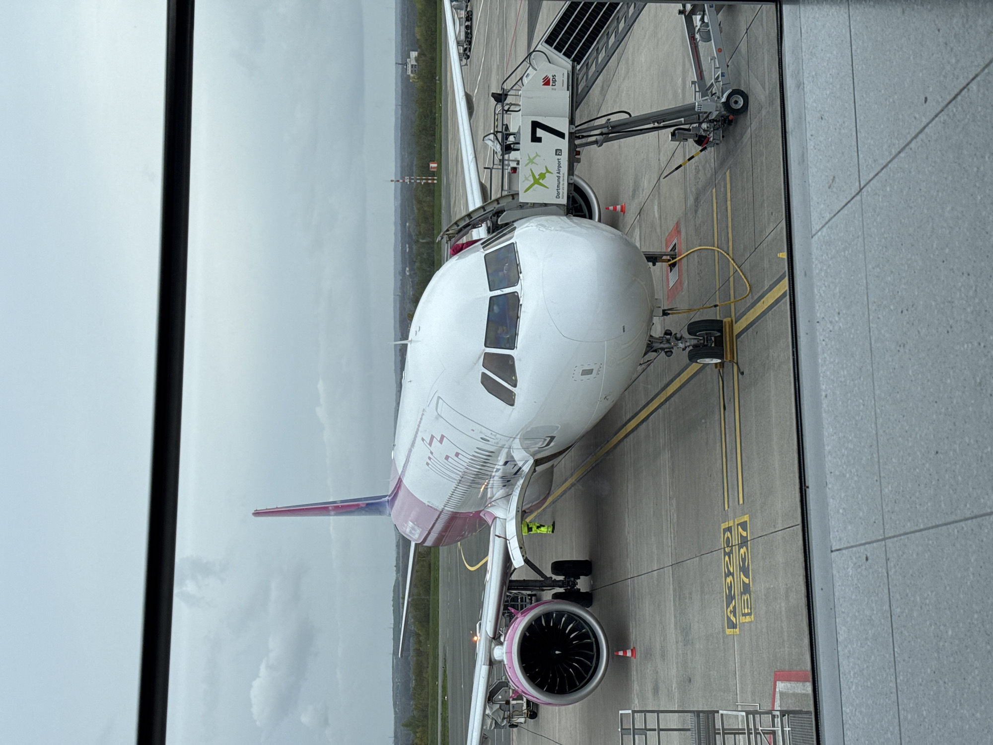 The image depicts a white and magenta Wizz Air Airbus A320 aircraft parked at gate 7 of Dortmund Airport (DTM) in Dortmund, Germany. A passenger boarding bridge is connected to the front of the aircraft. Two ground crew personnel are visible near the right engine. The weather appears overcast. The view is from inside the airport terminal, looking out through a window. The gate markings "A320 B737" are visible on the tarmac.