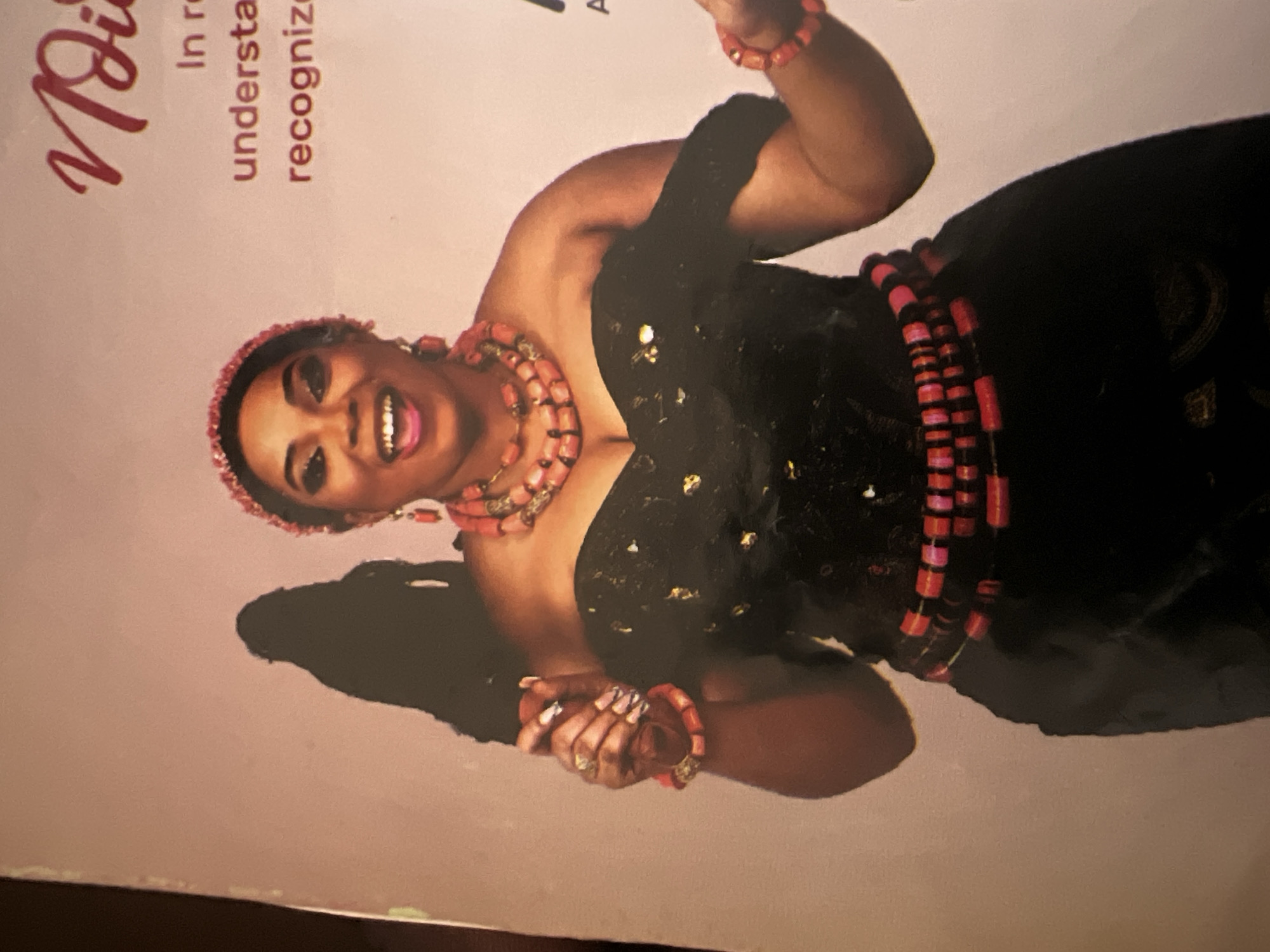 The image shows a woman in traditional Nigerian attire, likely from the Igbo ethnic group, given the coral bead jewelry. She is wearing a black off-the-shoulder dress adorned with gold accents, a coral bead necklace, earrings, bracelets, and a beaded belt. Her hair is styled up, and she has a beaded headband. She is smiling and appears to be in a celebratory mood. The background is a plain, light pink color with some text in a dark red font. The text appears to be a title and some descriptive phrases related to recognition or understanding. There is also a faint silhouette of another person or object behind the woman, to her left.
The image is a photograph, likely a studio portrait or a promotional image. There is no indication of the specific location within Jalingo, Nigeria, nor is there enough visual information to deduce the context of the image as it relates to the city itself. The scene is a close-up shot focusing on the woman and her attire.