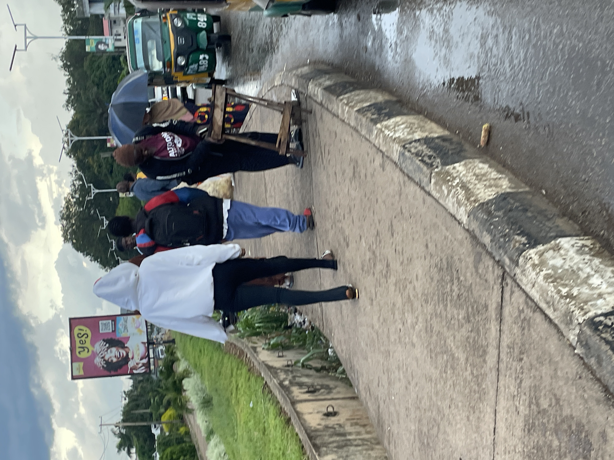 The image depicts a sidewalk scene in Barakin Lerawi, Nigeria, on a cloudy and wet day. A group of individuals are walking on the sidewalk. One person in the foreground wears a white hooded jacket and black leggings. Behind them, another person in a blue tracksuit carries a black backpack. Further back, two individuals are visible, one under a blue umbrella and another with a brown beanie and a dark jacket. To the right, a green and yellow auto-rickshaw with the license plate "P 14.83 840" is parked on the wet road. A wooden stand with items is positioned on the sidewalk. In the background, a billboard displays a smiling woman with the text "Yes!". The environment suggests a typical street scene with a sidewalk, road, vegetation, and utility poles.