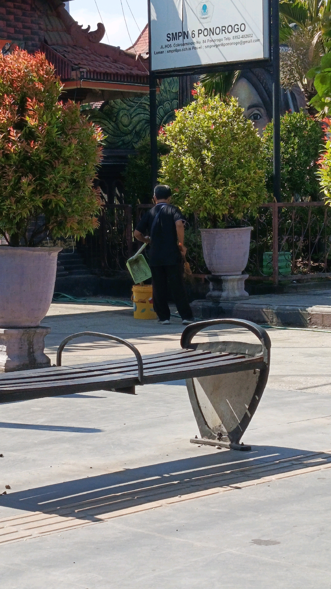 A man in a black t-shirt and pants is cleaning the paved area in front of a building. He is holding a green dustpan and a broom, and a yellow bucket is on the ground near him. Behind him, there are large potted plants with red and green foliage, and a building with a red tiled roof. A sign that reads "SMPN 6 PONOROGO" is visible above the building. The scene is illuminated by sunlight, casting shadows on the ground. The location appears to be a school campus in Ponorogo, Indonesia.