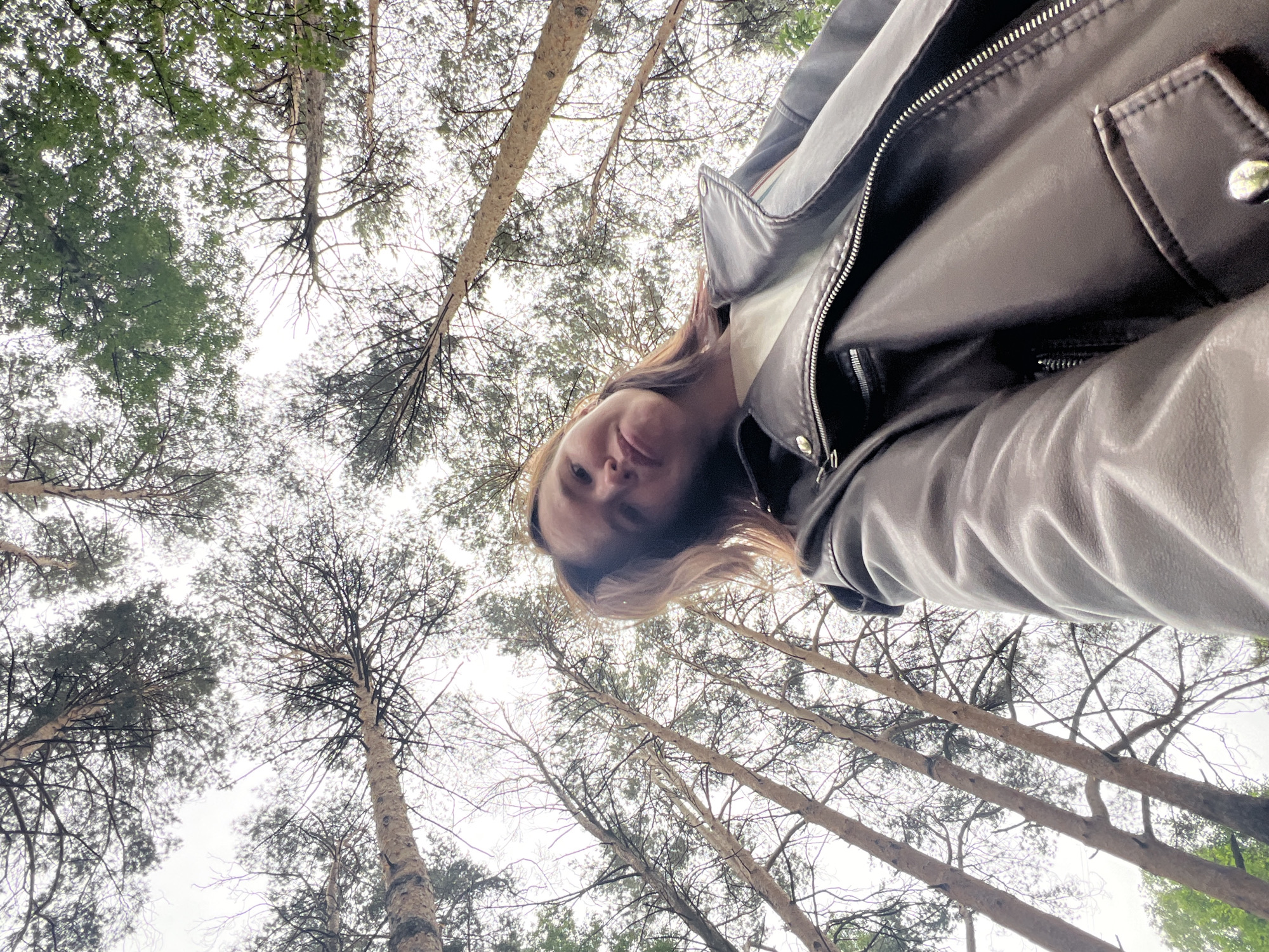 A low-angle shot depicts a person wearing a brown leather jacket, looking down at the camera. The background consists of tall trees with sparse foliage reaching towards a cloudy sky. The photograph appears to have been taken in a wooded area. Given the prompt's request, this likely took place in Chaikovsky, Russia.