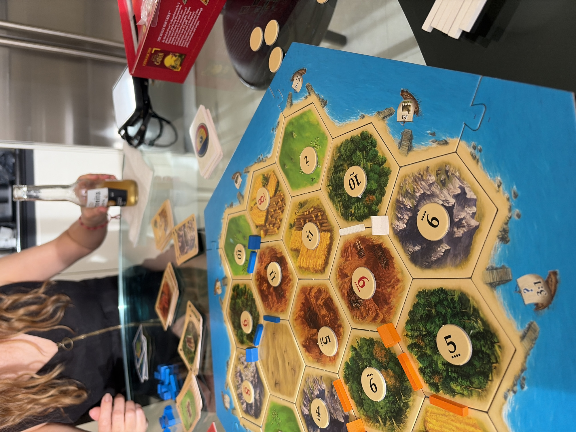 The image captures a game of "The Rivals for Catan" being played on a glass table. The game board, composed of hexagonal tiles representing different terrains, is in the foreground. Various numbered tokens and resource cards are scattered around the board. Game pieces in blue and orange are placed on the board, indicating player positions. A Corona beer bottle rests on the table's surface, near a player's hand. A game box and dice are visible in the background. The reflection in the glass reveals part of a kitchen setting, including stainless steel cabinets.
While the image provides context, it lacks specific details to definitively establish its location as Key Biscayne, United States of America. The scene depicts a domestic setting related to a board game and beverage.