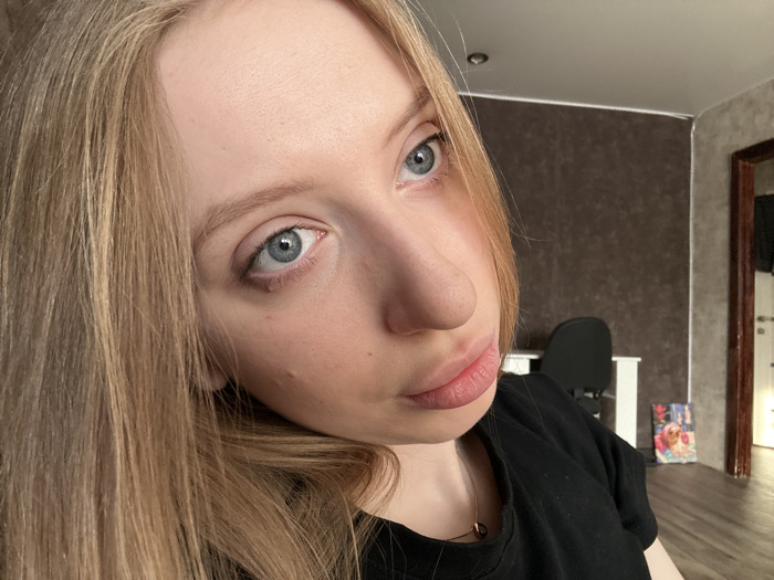 A close-up, eye-level selfie-style photograph captures a young woman indoors in what appears to be a residential room in the city of Chaikovsky, Russia. The woman, identified as the primary subject, has fair skin, light blue eyes, and long, straight, blonde hair that cascades over her shoulders. Her gaze is directed straight into the camera. She wears a black t-shirt and a thin, gold-colored necklace with a small, round pendant. Her lips are slightly parted, and her natural eyebrows are light blonde. Two small moles or blemishes are visible on her right cheek (viewer's left).
The background environment consists of interior walls, a ceiling, and a floor. The wall behind her left shoulder (viewer's right) is textured and dark brown, while the wall to her right (viewer's left) is lighter, appearing off-white or light gray. A plain white ceiling is visible at the top, featuring a small, dark, circular fixture near the upper left corner. The floor, visible towards the bottom right, is dark wood or laminate. Furniture in the background includes a partial view of a black office chair and a white desk or table surface. A colorful framed picture or poster leans against the dark brown wall on the floor. To the far right, a brown wooden door frame opens into another room with a partially visible light-colored door.