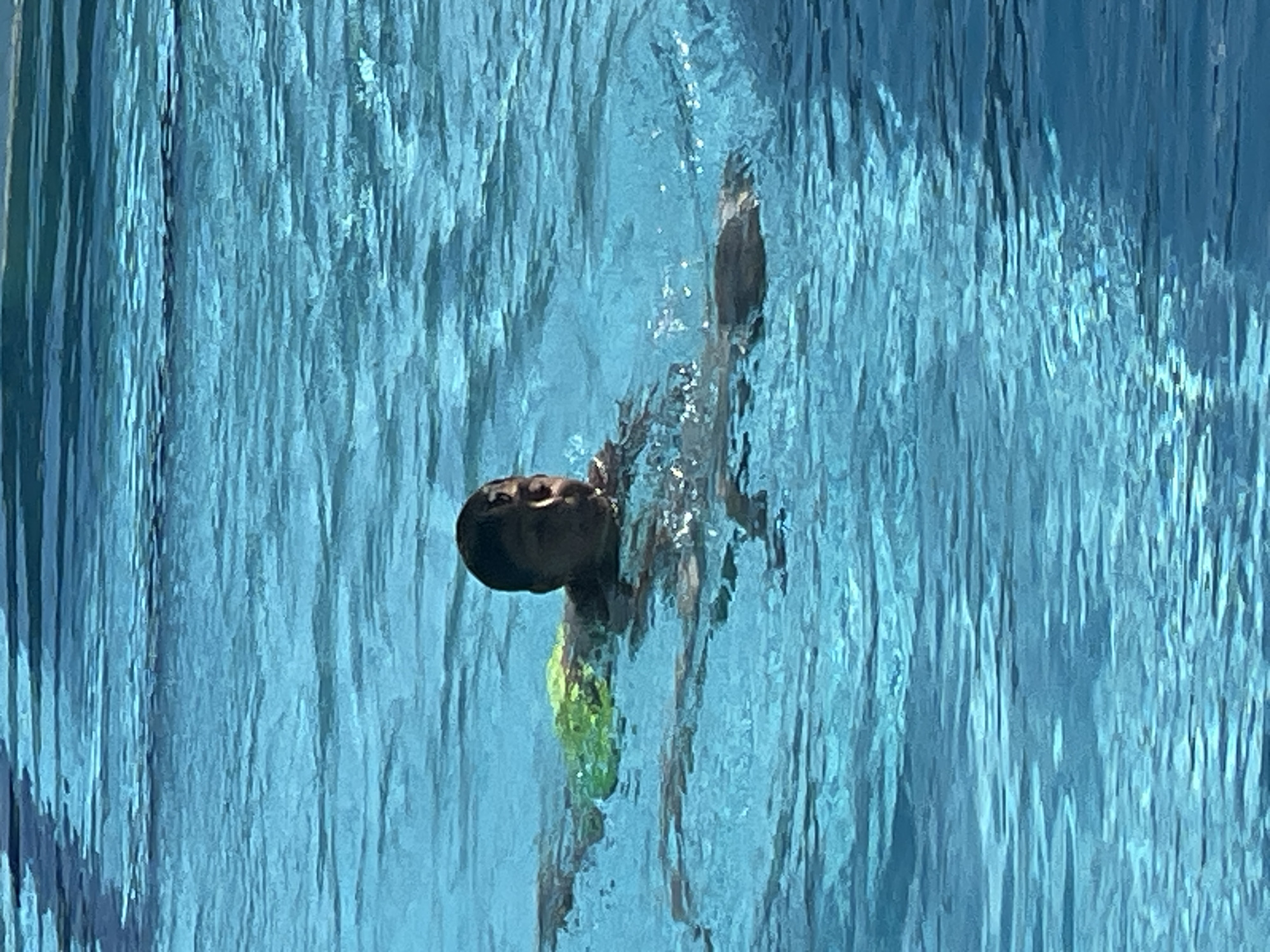 A child is swimming in a bright blue swimming pool. The child is of dark complexion and is wearing green swim trunks. The child's head is above the water and they are looking to the right of the frame. The water is choppy, indicating movement. Sunlight reflects off the surface of the water, creating bright patches. The background is entirely composed of the blue water of the pool, with some subtle variations in color and texture due to ripples and light.