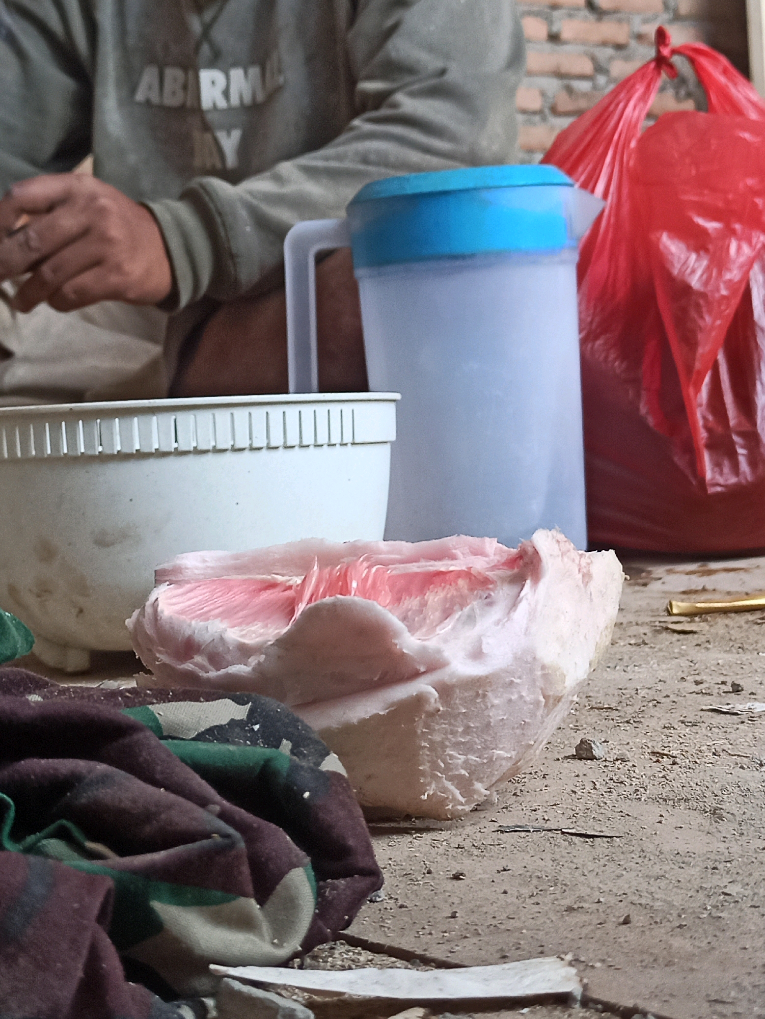 An outdoor, close-up shot depicts a portion of a person sitting in the foreground, partially obscured by camouflage fabric and a white bowl. The person wears a grey sweatshirt with white lettering that reads "ABIRMA Y". Their hands are visible, holding what appears to be a cigarette. To the right of the person, a clear plastic pitcher with a blue lid is positioned next to a crinkled red plastic bag. A piece of pale pink fruit, possibly a grapefruit or pomelo, rests on the camouflage fabric in front of the white bowl. The ground is composed of dirt and debris, with a few small stones scattered about. In the background, a brick wall is visible. The lighting is dim, suggesting an overcast day or shaded area.