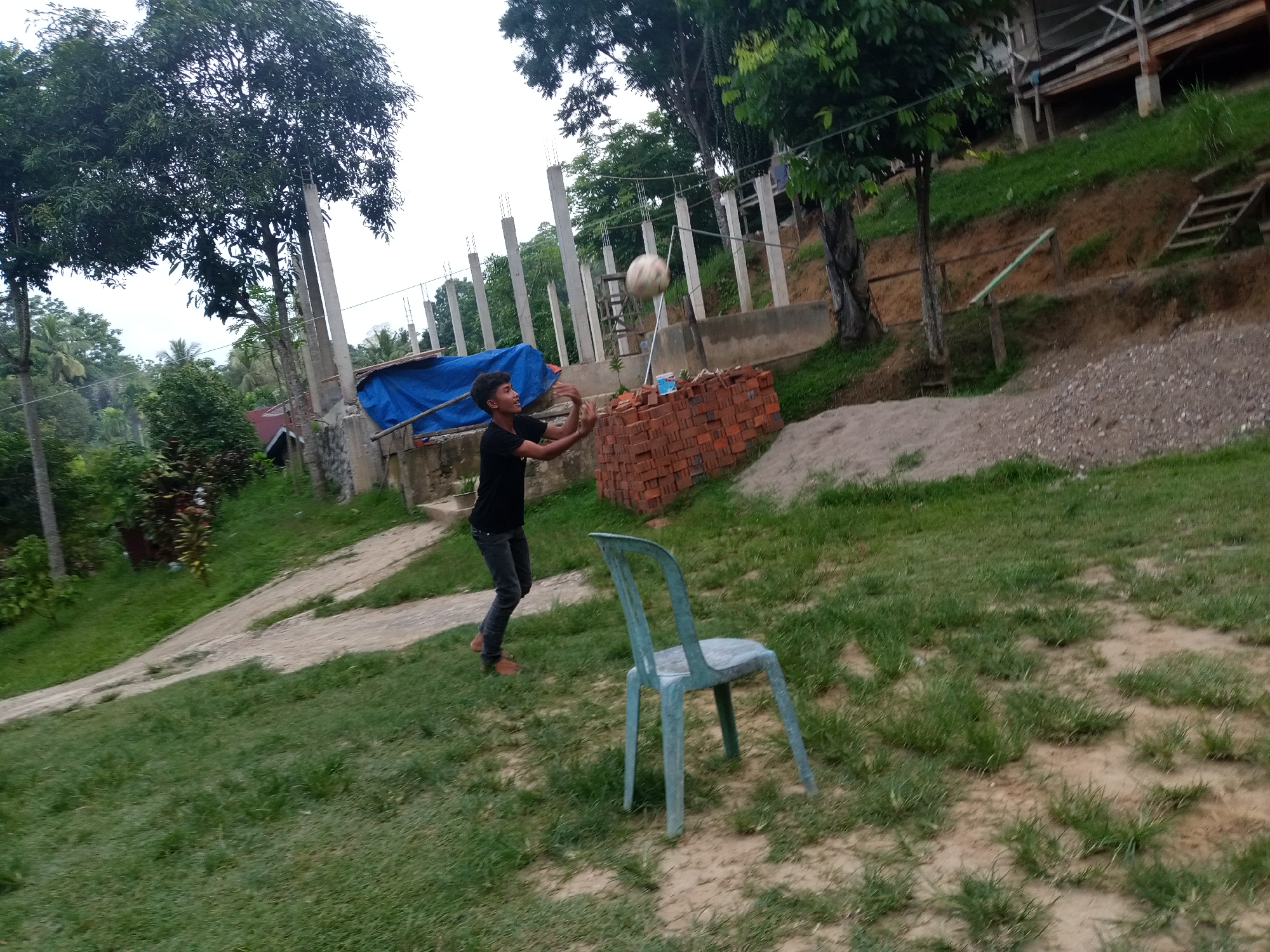 A young boy is playing with a ball in an outdoor setting, likely Lhokseumawe, Indonesia. He is wearing a black t-shirt and dark pants, with brown sandals. The boy is mid-action, tossing the ball upwards with his hands. He appears focused and engaged in the game.
The scene is set on a grassy area with a dirt path winding through it. In the background, there are construction elements, including concrete pillars and what appears to be a blue tarp covering some materials. A stack of red bricks is visible near the boy, and a light blue plastic chair sits on the grass to his right. Trees with lush green foliage are scattered around the area, and a building structure can be seen in the upper right portion of the image, suggesting it is an elevated structure. The ground is uneven, with some areas of exposed dirt and gravel. The lighting suggests it is daytime, and the weather appears to be overcast or hazy. The overall impression is a casual, outdoor activity taking place in a developing or unfinished area.