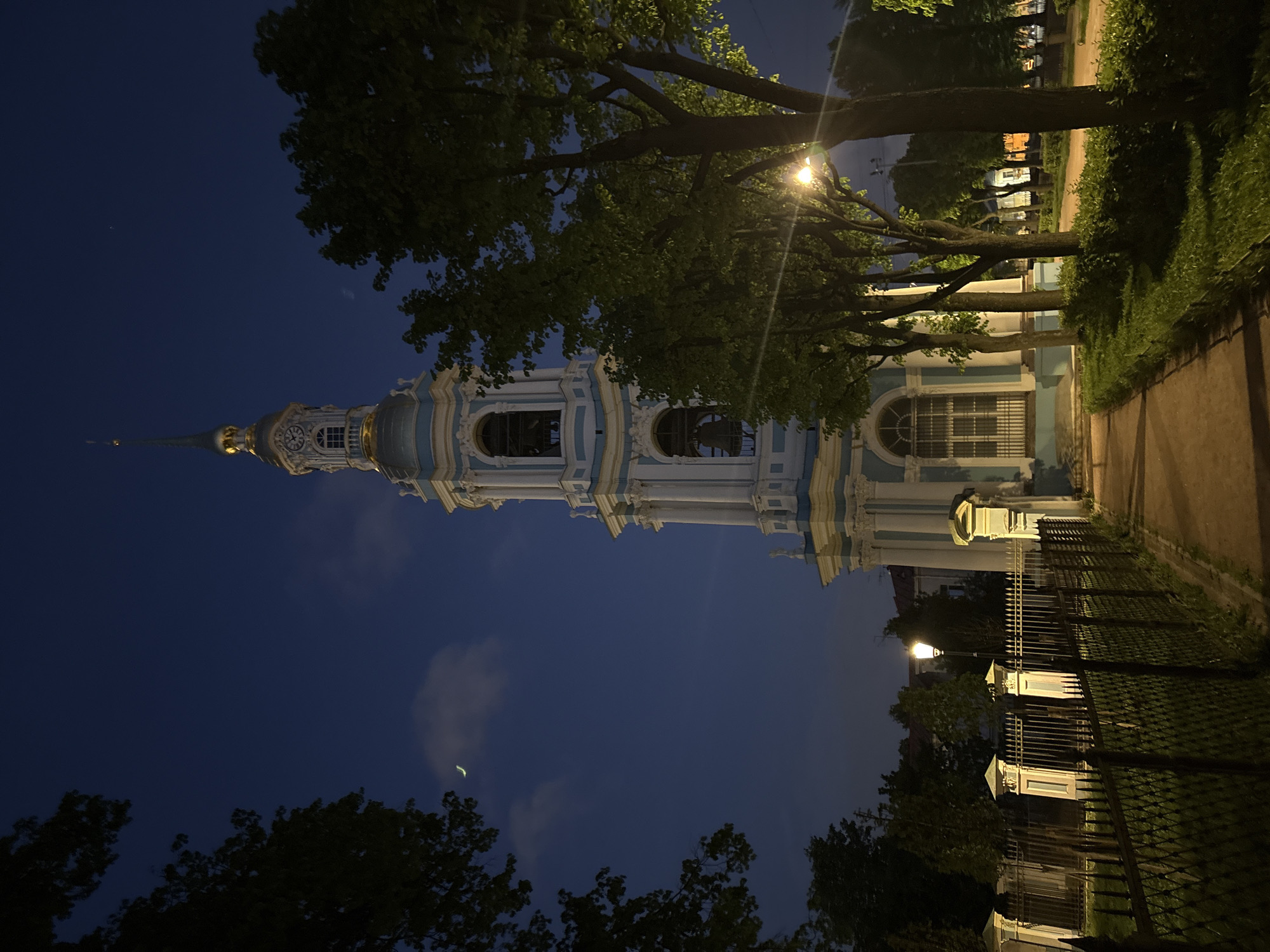 The image shows St. Nicholas Naval Cathedral in St. Petersburg, Russia, at night. The cathedral, with its white and blue baroque facade and tall, gilded spire, dominates the frame. The sky is a dark blue, with a sliver of a crescent moon visible. Trees frame the cathedral, some obscuring parts of the building and sky. A street lamp illuminates the surrounding area. A fence lines the right side of the frame along a walkway.