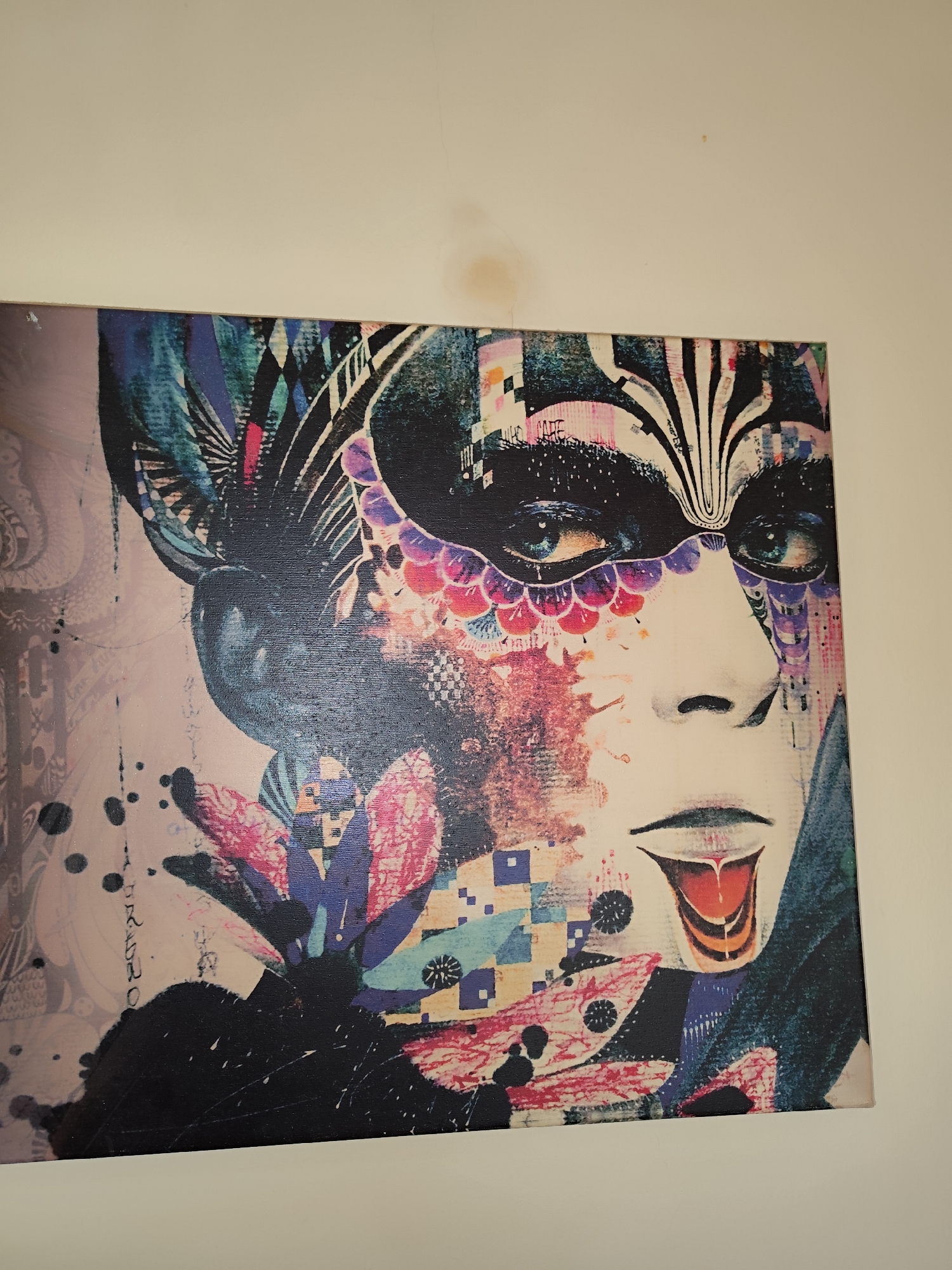The image shows a portrait of a person wearing a mask. The mask is decorated with various colors and patterns, including floral and geometric designs. The person's face is painted in a pale color with red paint at the mouth, appearing as if the person's tongue is dripping. The background of the portrait is composed of abstract patterns and colors. The image appears to be a photograph of a printed artwork hanging on a white wall. The location in Sarıyer, Turkey is not directly discernible from the image.