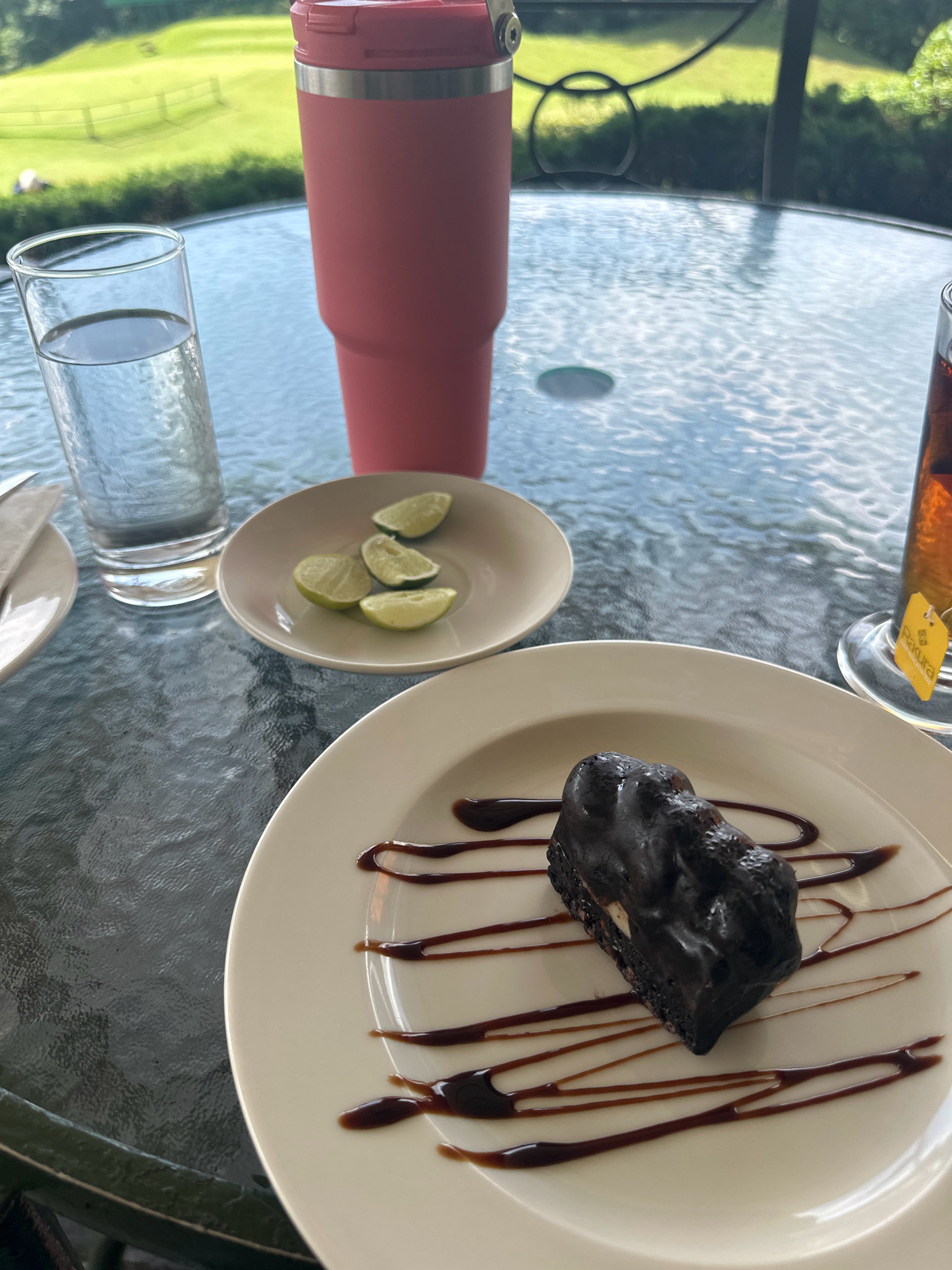 A close-up shot captures a dessert and drinks on a glass table outdoors, with a blurred background of a golf course and green foliage. In the foreground, a white plate holds a slice of dark chocolate cake, drizzled with chocolate sauce. To the left, a clear glass of water sits next to a small white dish containing four lime wedges. Behind these, a tall, pink insulated tumbler stands prominently. To the right, a glass of amber liquid, likely tea, with a yellow tag attached to a string is visible. The table's surface reflects the sky and surrounding greenery. The setting suggests a leisurely break or meal in an outdoor cafe or restaurant. This scene is located in Kageshwori Manohara, Nepal.