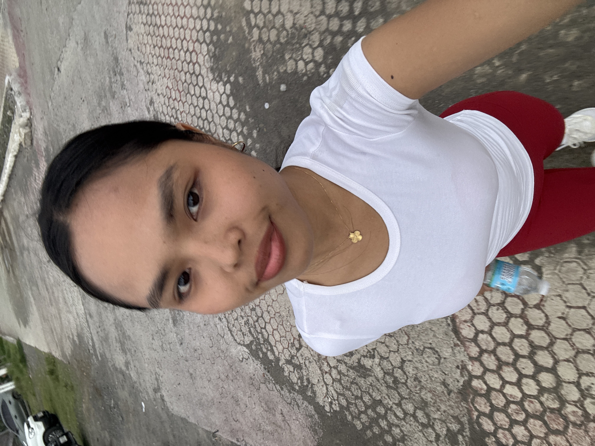 An outdoor selfie features a young woman looking directly at the camera, taken from a slightly high angle. She has dark hair pulled back and wears a white short-sleeved t-shirt, red athletic pants, and white sneakers. A delicate gold necklace with a small, gold four-leaf clover or flower-shaped charm is visible around her neck, along with a small gold hoop earring on her right ear.
The predominant background is a concrete ground exhibiting a distinct hexagonal or honeycomb pattern, which appears somewhat worn in areas. In the far left background, parts of a white car and some green grass are visible, suggesting an urban or suburban setting. A clear plastic water bottle with a blue label, potentially reading "SPRING" or a similar brand, lies on the ground near her right leg. The lighting is soft and diffused, indicative of an overcast day. The woman has a neutral expression with a slight hint of a smile. The scene is set in Davao City, Philippines.