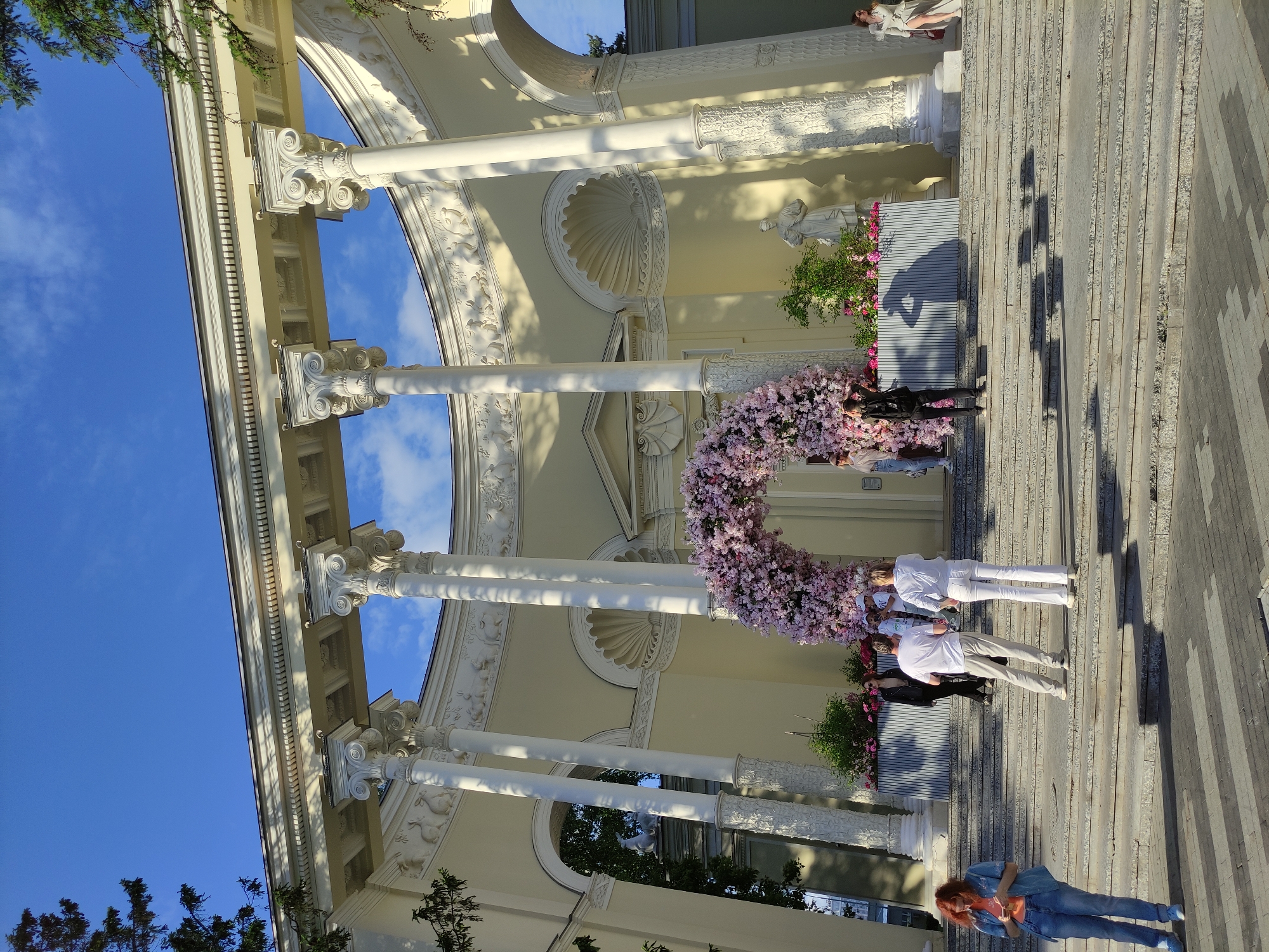 The image depicts a scene at VDNKh, a park and exhibition center in Moscow, Russia. The architecture features classical columns and ornate decorations, possibly part of a pavilion or entrance structure. A floral arrangement, predominantly pink, forms a semi-circular arch. Several people are present: one woman is wearing a blue jacket and jeans; two people are dressed in white outfits; others are standing in the background. The scene appears to be a public space, with stone steps leading up to the structure. The sky is visible through the columns.