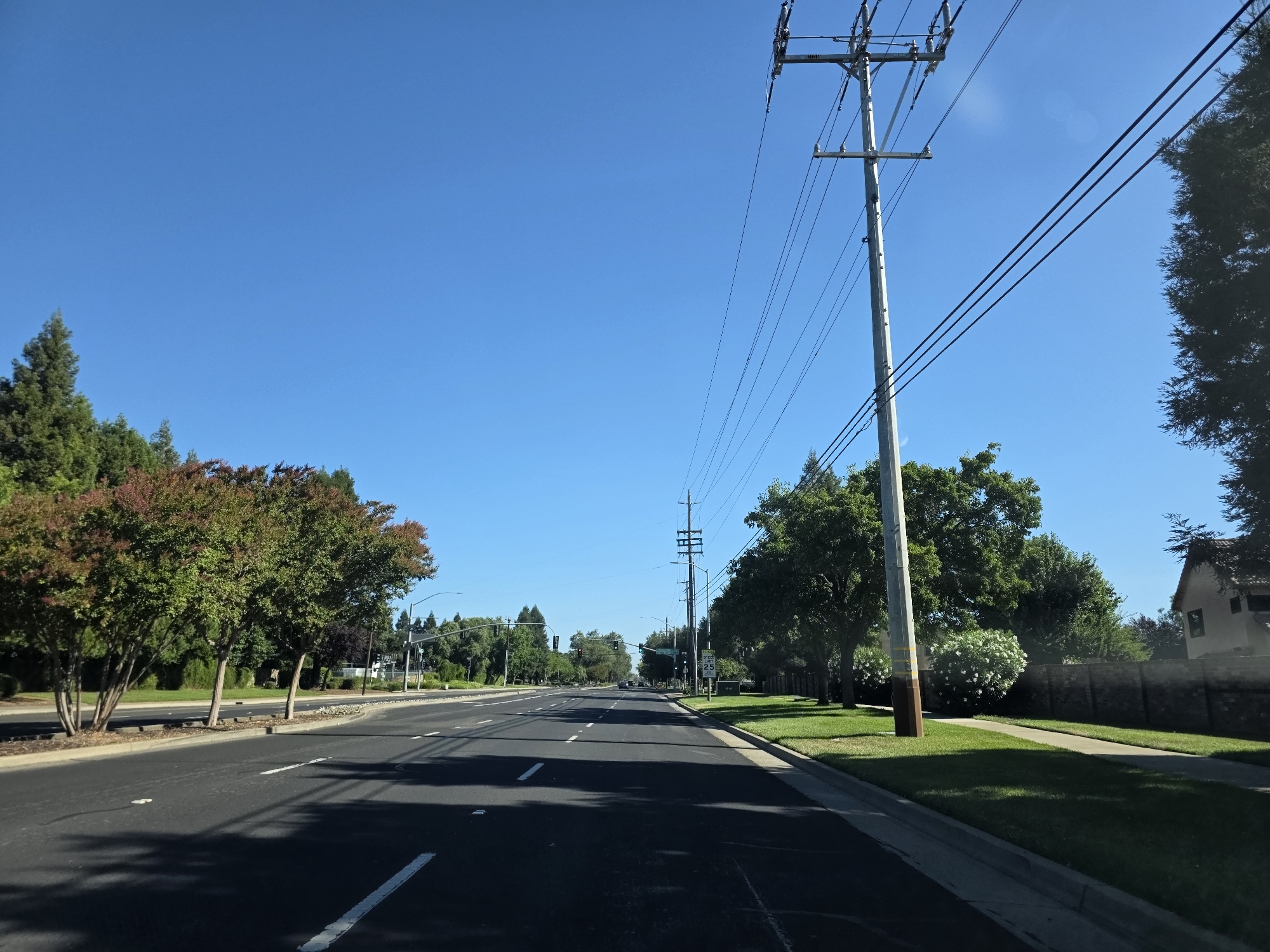 The image depicts a clear, sunny day with a bright blue sky over a street in Elk Grove, California, United States of America. The perspective is from a vehicle traveling down a multi-lane road, suggesting an urban or suburban setting.
On the left side of the road, a row of trees with green and reddish foliage lines the sidewalk and a well-maintained lawn. The trees appear to be deciduous, possibly in the process of changing colors. The road itself is asphalt with visible white lane markers and some scattered white dots.
On the right side of the road, a prominent utility pole with multiple power lines stretches diagonally across the frame. Beyond the pole, there is a residential area with a brick wall bordering a manicured lawn and a house partially visible. More trees, including a large evergreen, are situated in this area.
In the distance, the road continues with more trees and streetlights visible. Traffic signals are present, and a speed limit sign indicating "25" is mounted on a pole. The overall scene is one of a typical suburban street on a pleasant day. There are no people or animals visible in the image.