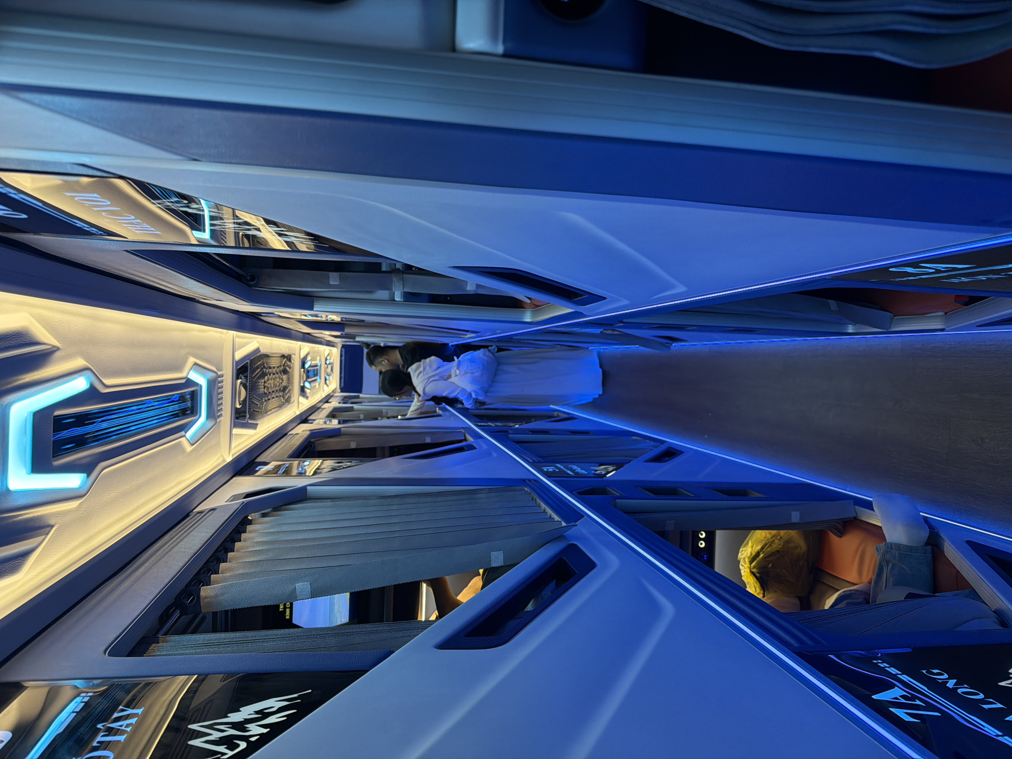 The image depicts the interior of a modern sleeper bus, oriented as if looking down a narrow aisle between individual sleeping berths. The design is sleek and futuristic, featuring a dominant color palette of blue and white, accented by both cool blue and warm yellow LED strip lighting.
Several people are visible within the bus. In the mid-ground, a person wearing a white shirt and dark pants stands or leans in the aisle. Further down, another individual in a white garment is present. Towards the foreground on the right, a person with a yellow covering over their head is seen resting inside one of the berths, suggesting a state of travel or relaxation.
The bus is equipped with capsule-like berths on both sides of the aisle, likely a double-decker configuration. Each berth appears to have a privacy screen or window blind, one of which is partially open. The interior surfaces are reflective, mirroring the neon blue light strips and other elements of the cabin. The overall atmosphere is calm, lit by the artificial glow of the interior lighting, suggesting travel during evening or nighttime hours.
Visible text on the berths and reflective surfaces includes "7A," likely a berth number, and "HA LONG," indicating Hạ Long, a popular destination in Vietnam. Other partially visible text includes "TÂY" on the left and "KÍNH CH" on a blind, which could be part of "KÍNH CHÚC" (wishing you) or related to a screen.