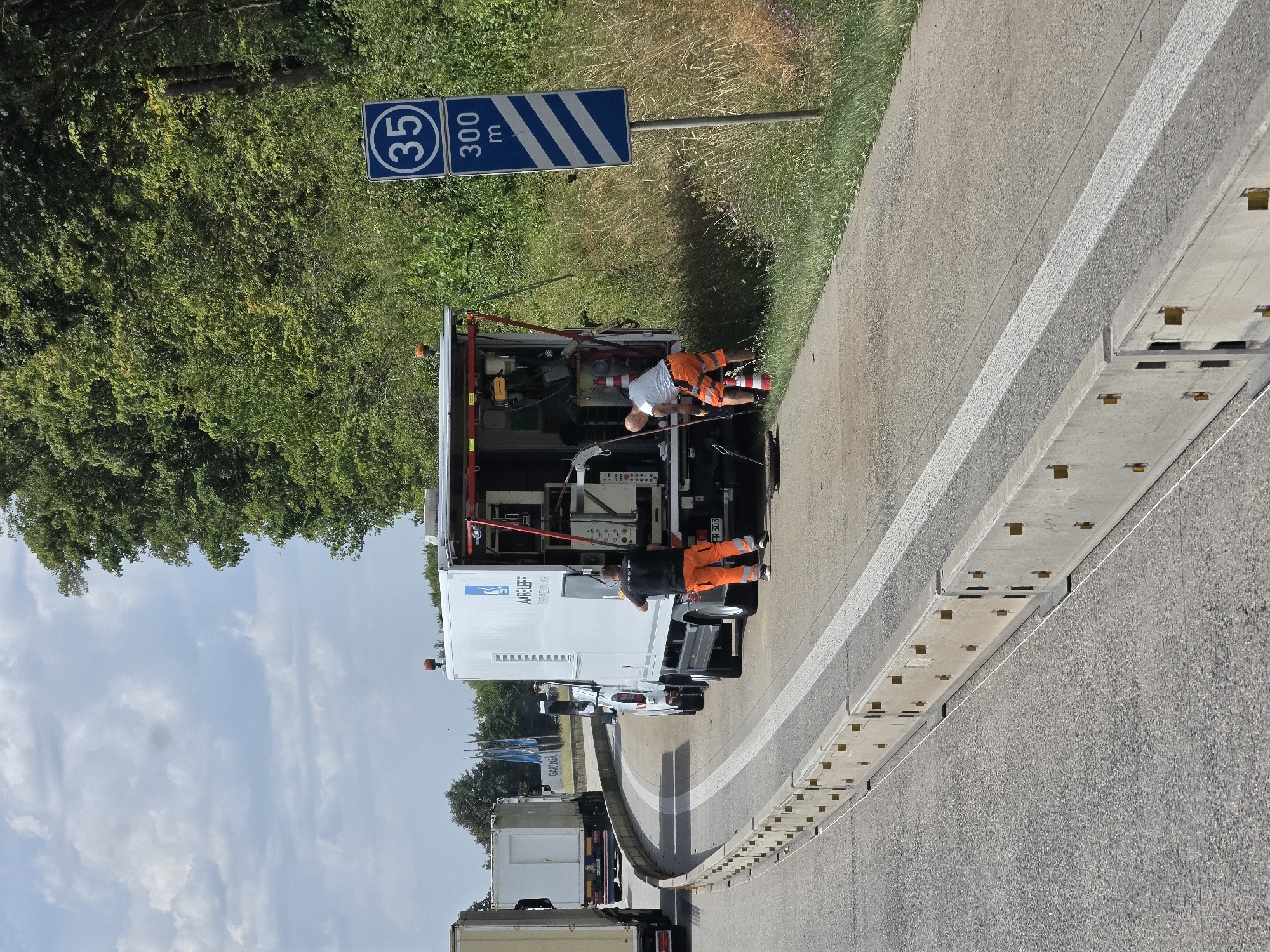 In Neustadt (Wied), Germany, on a highway, two individuals are observed working next to a white truck with its rear open, revealing machinery. One person, wearing a black shirt and orange pants, stands near the back of the truck, facing away from the camera. The other person, in a white shirt and orange shorts, is bent over near a manhole cover, holding a long tool. A white van is parked in front of the truck, and another truck is visible to the left. A road sign indicating "35 300 m" with diagonal stripes is positioned on the right side of the image. The scene takes place on a paved road with concrete barriers and white lane markings, bordered by a grassy embankment and dense green trees. The sky is partly cloudy.