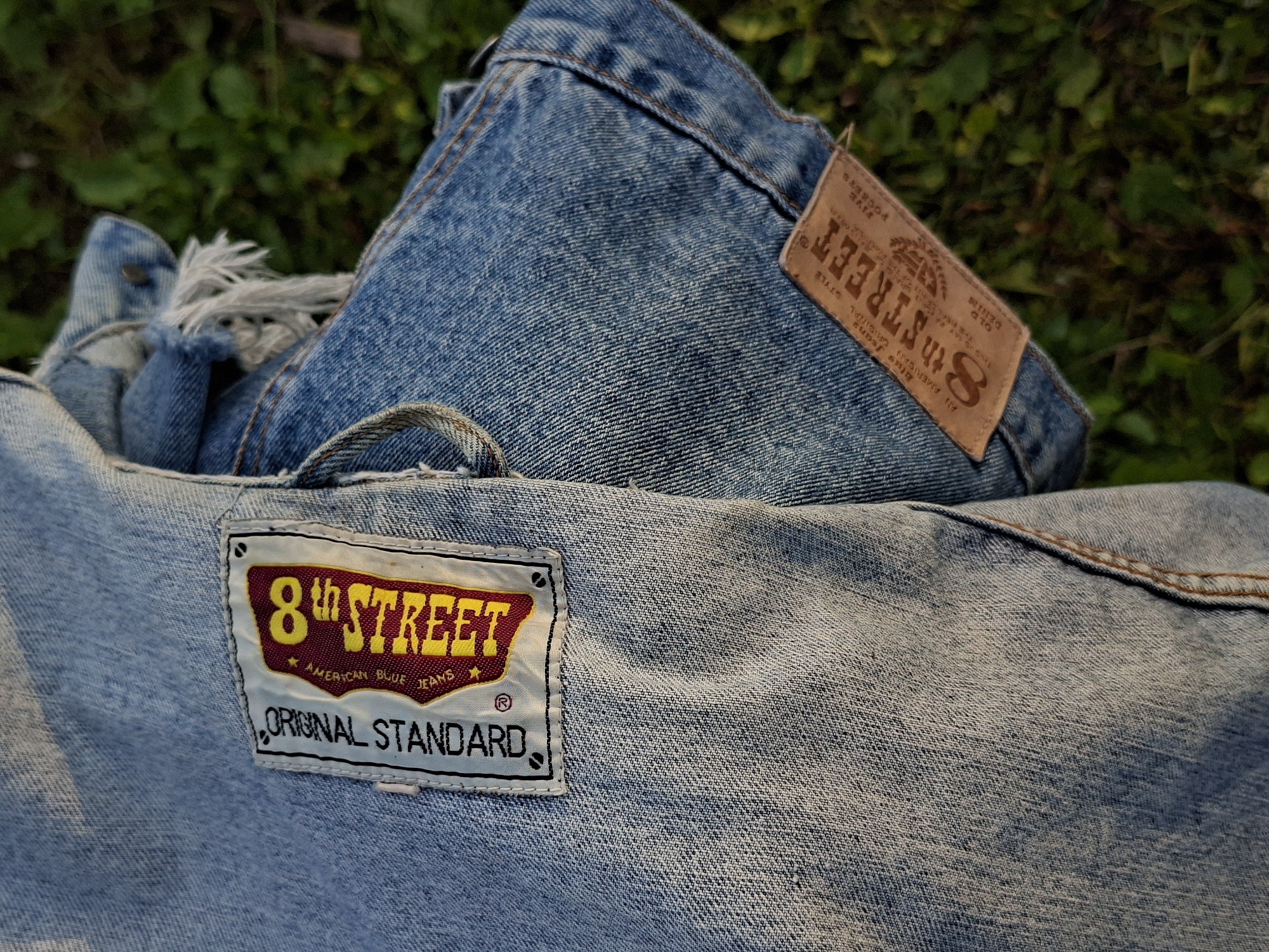The image shows two pieces of denim fabric, likely from jeans or a jacket, lying on a patch of green grass. One piece features a label with the text "8th STREET" in yellow and red lettering, along with "ORIGINAL STANDARD" and smaller text that is difficult to read. The other piece has a brown leather tag with "8th STREET" and other text. Both pieces show signs of wear, with frayed edges and a weathered appearance. The background is a blur of green foliage, suggesting an outdoor setting. There are no people or specific landmarks visible in the image.